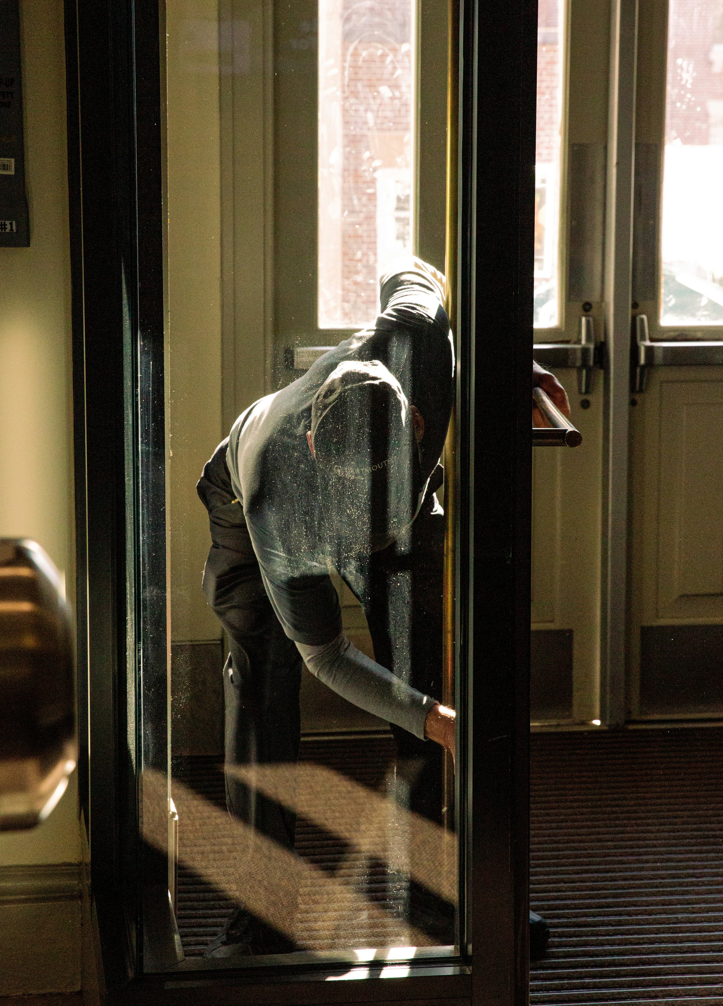 A person cleaning a glass door with their arm extended inside a building, viewed from outside.