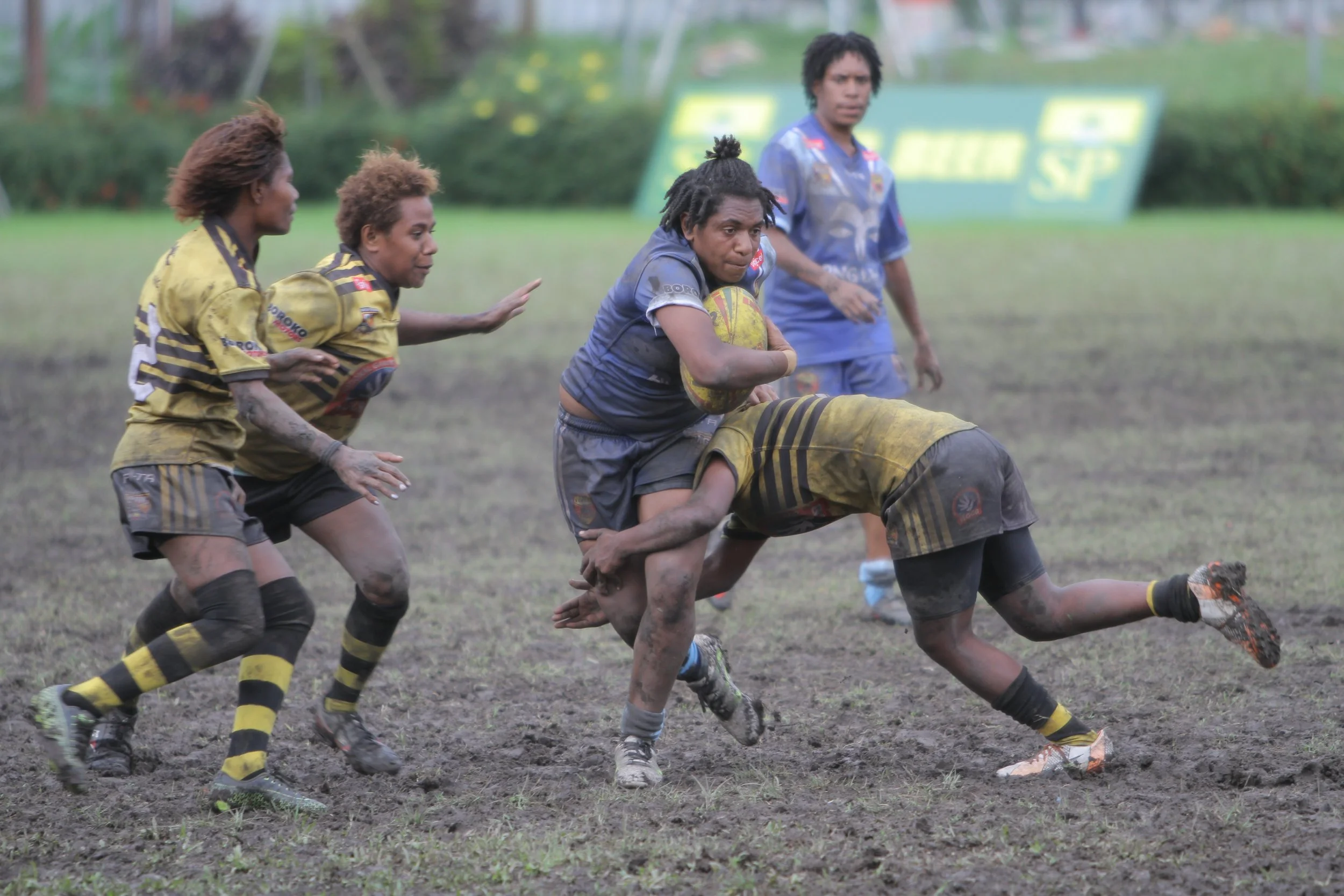 Female rugby league players in muddy field, with some wearing yellow and black uniforms, competing for the ball during a game.