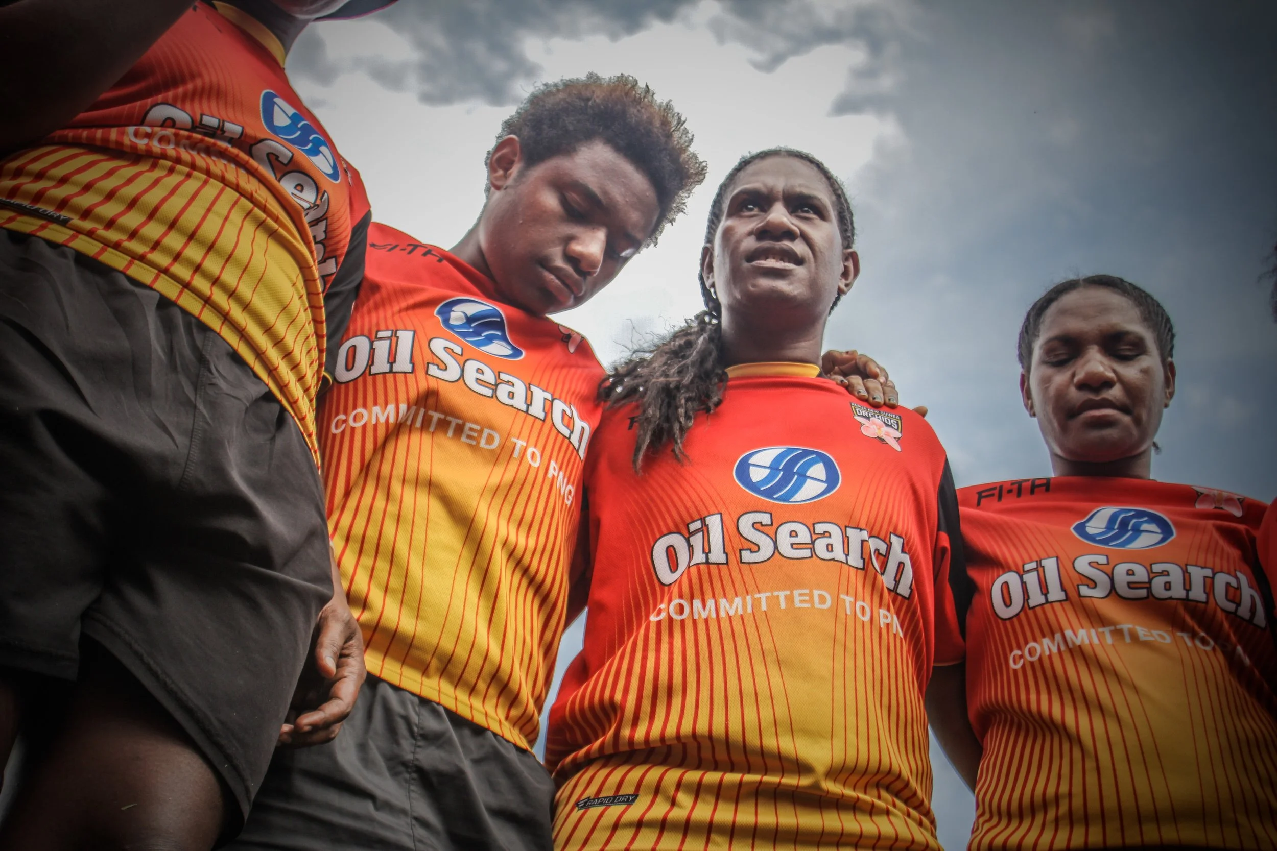 Four female rugby league players standing in a line, wearing orange jerseys with sponsorship logos, under a cloudy sky. They have serious expressions and are looking down at the camera.