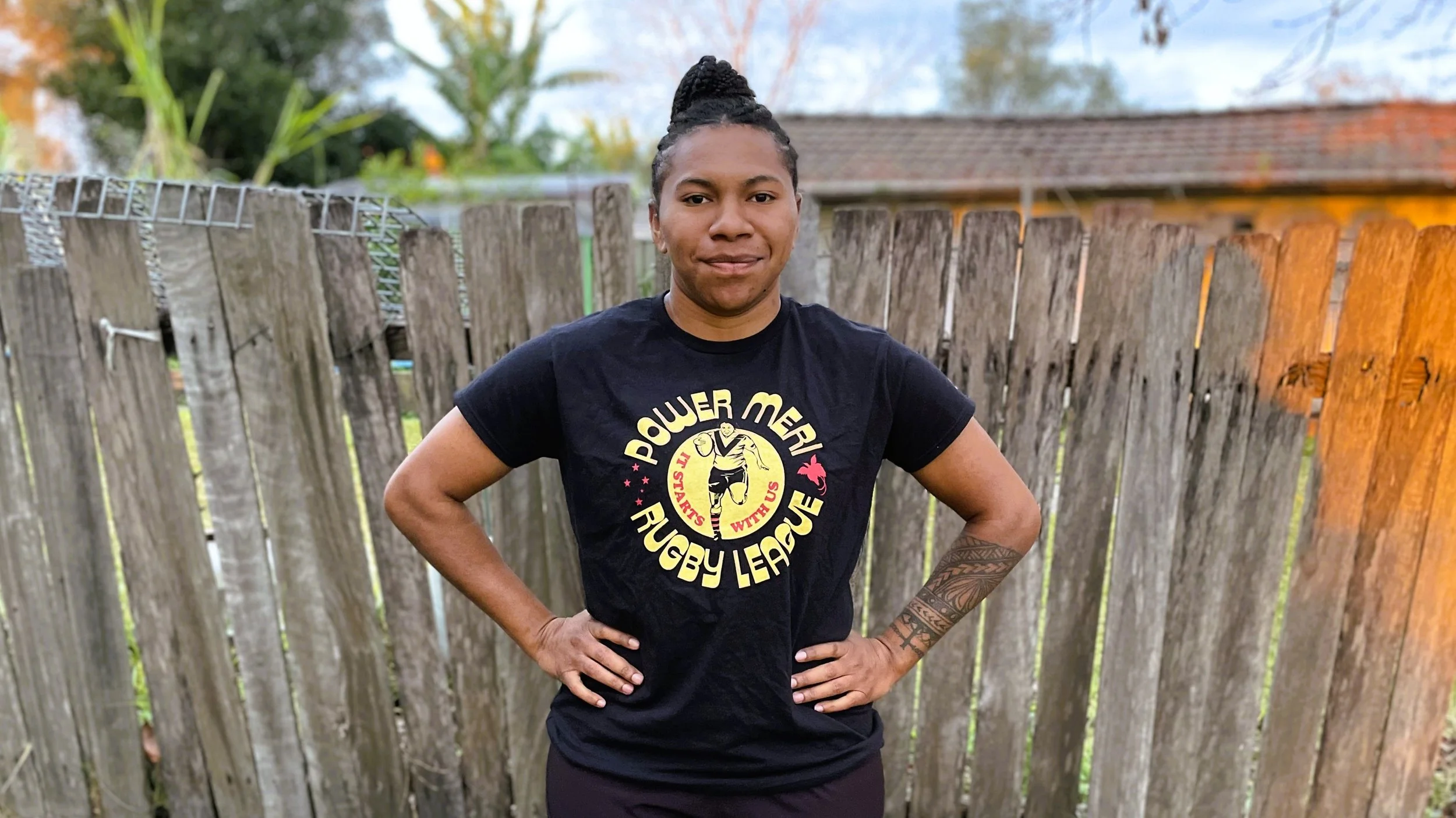A woman standing outdoors in front of a wooden fence with her hands on her hips, wearing a black t-shirt with 'Rugby League' and a graphic of a rugby player, with trees and a house in the background.