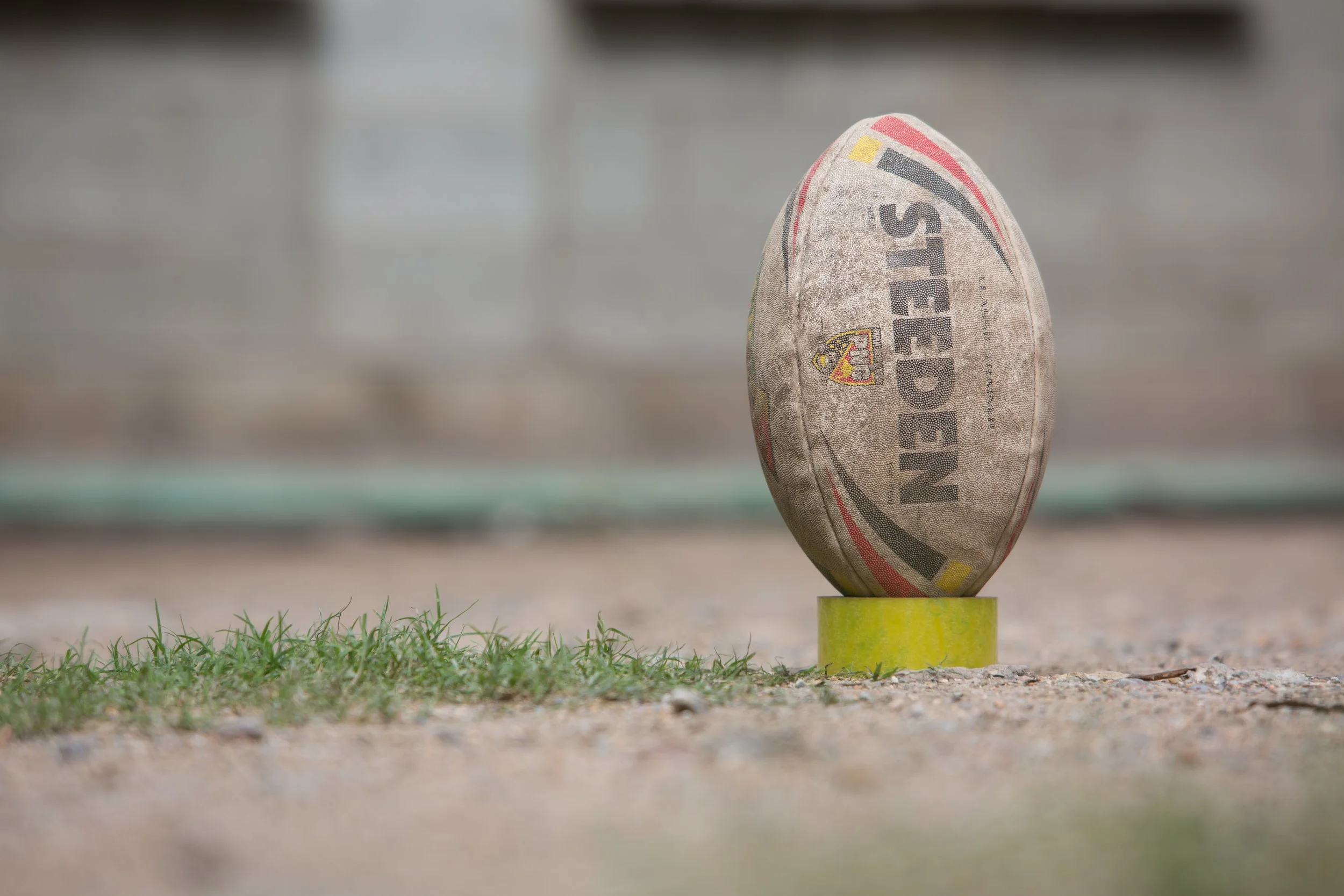 A rugby league ball with a worn, dirty appearance stands upright on a sticky tape roll in place of a kicking tee on a patch of grass, with a blurred background of pavement or a running track.