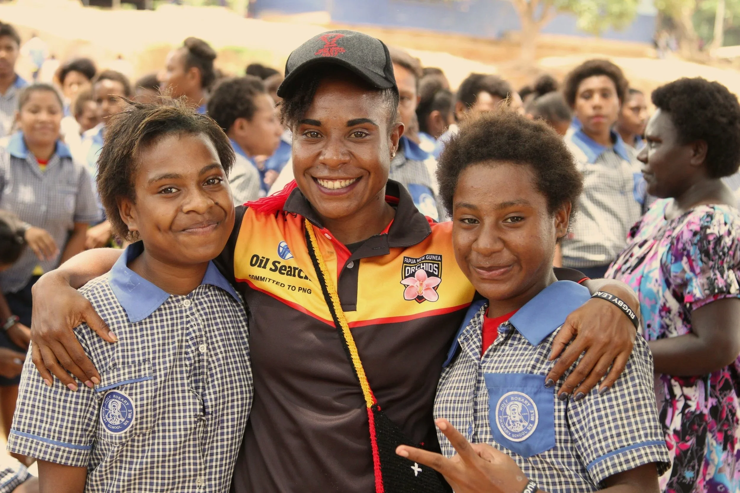 A smiling female rugby player wearing a black cap and a sports jersey, surrounded by young girls in school uniforms, posing for a group photo outdoors.