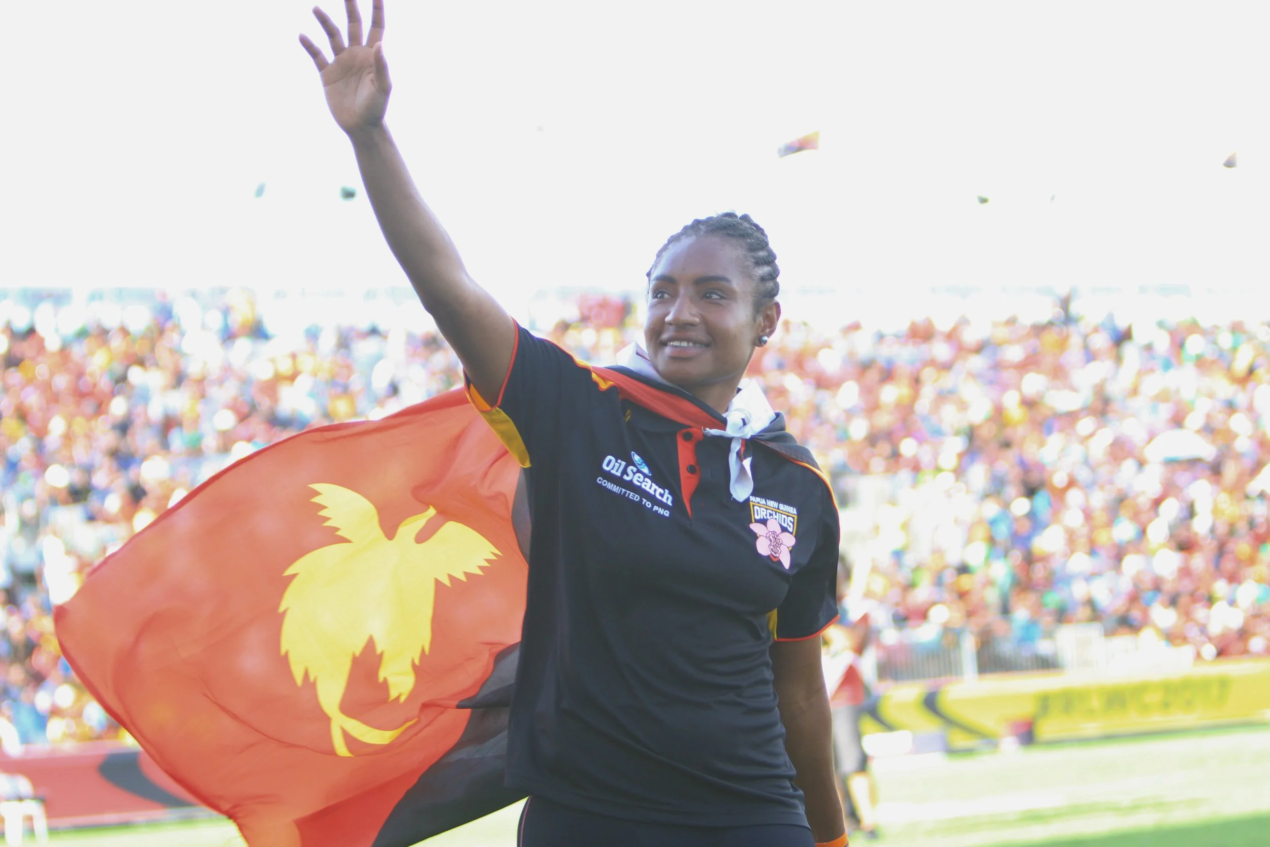 A female rugby league player waving with a smile, wearing a black and red jersey, carrying an Papua New Guinea flag in a stadium filled with spectators.