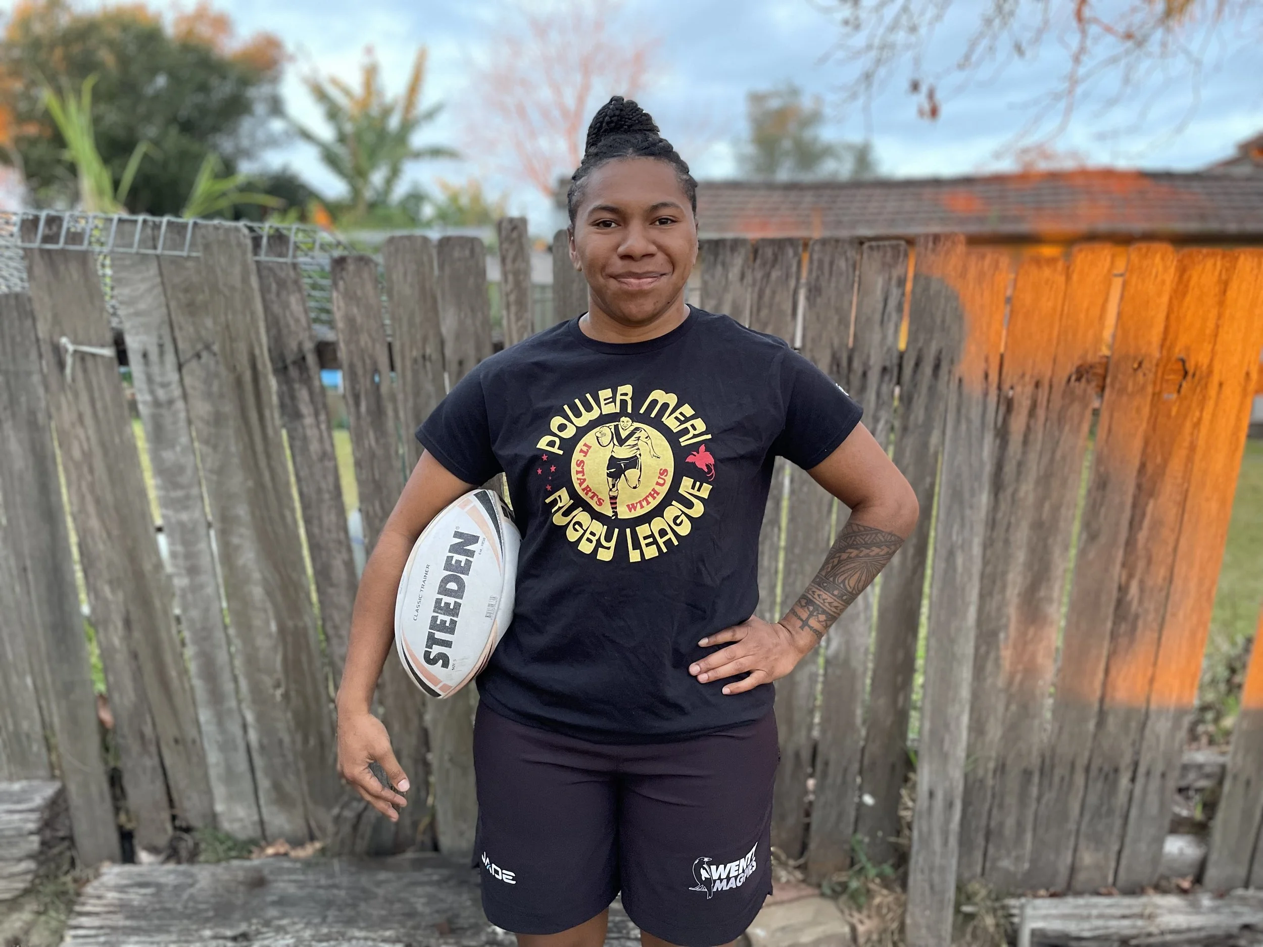 Woman standing outdoors near a wooden fence, holding a rugby ball, wearing a black t-shirt with 'Rugby League', with trees and a cloudy sky in the background.