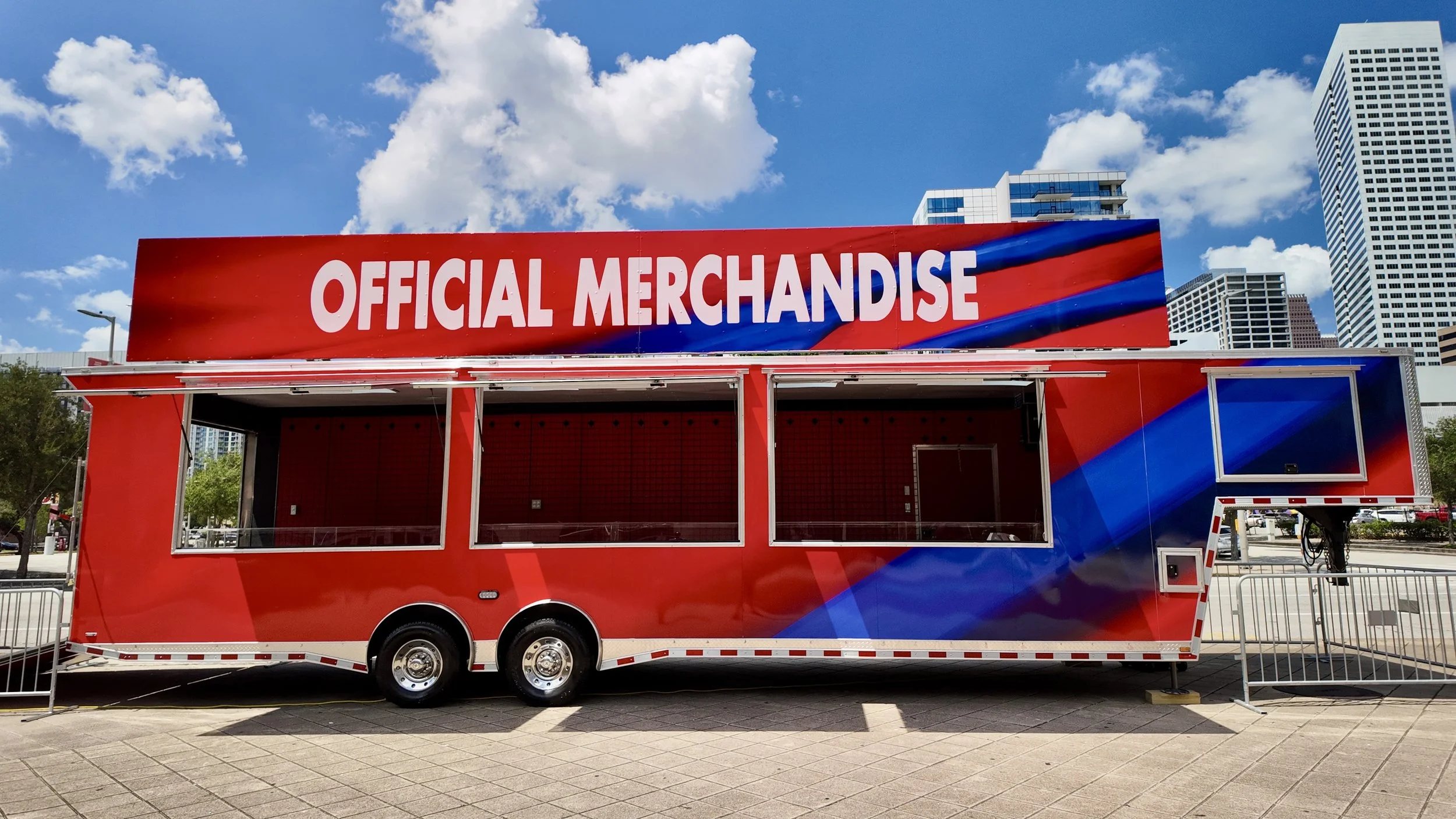 Red promotional trailer with a large sign that says 'Official Merchandise' parked outdoors in an urban area with skyscrapers and blue skies in the background.