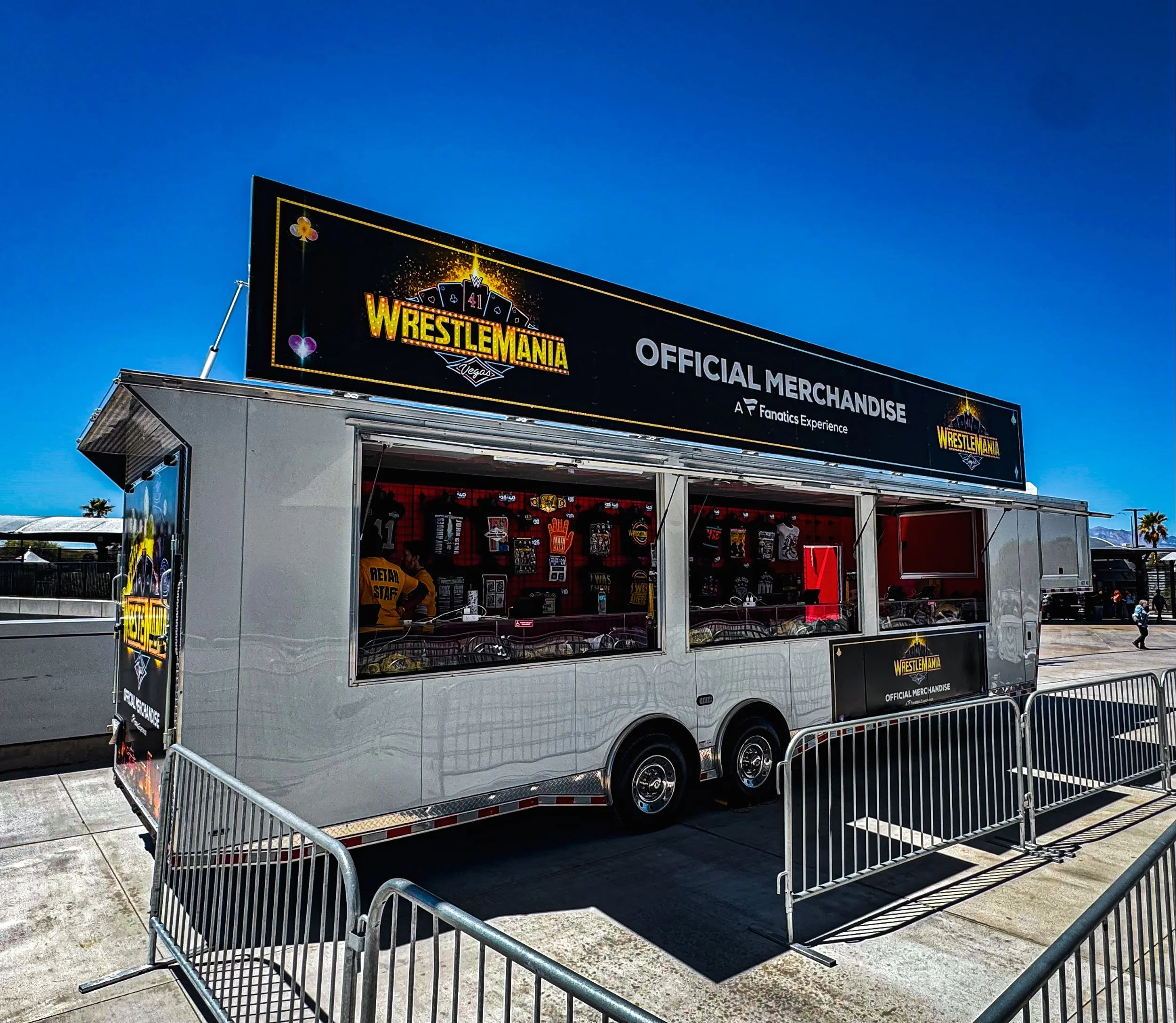 WrestleMania official merchandise trailer at an outdoor event under a clear blue sky, with metal barricades in front.