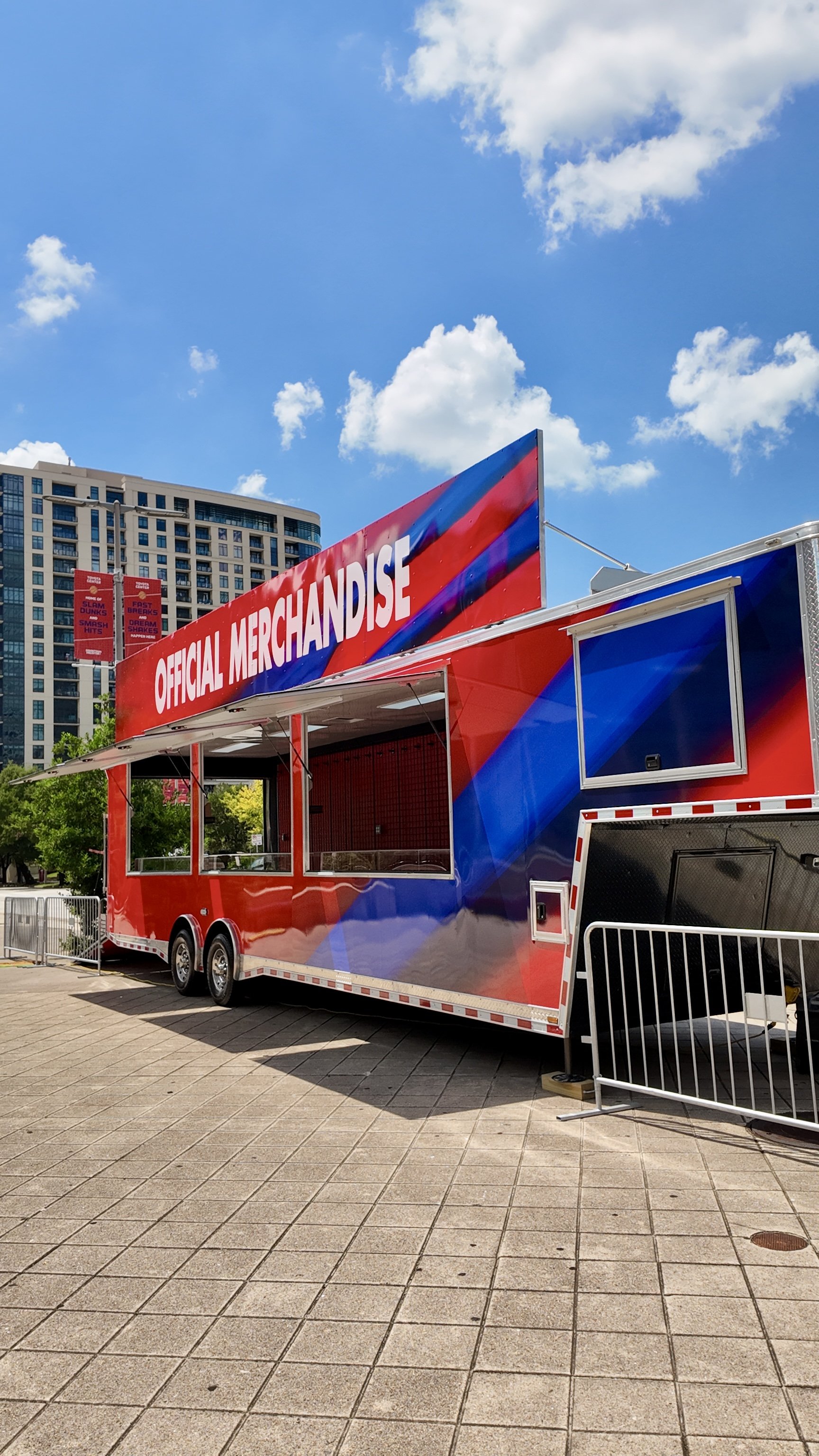 A red, blue, and white merchandise trailer with an open window and a sign that says 'Official Merchandise' on top, set up on a paved outdoor area with a multi-story building and trees in the background under a partly cloudy sky.