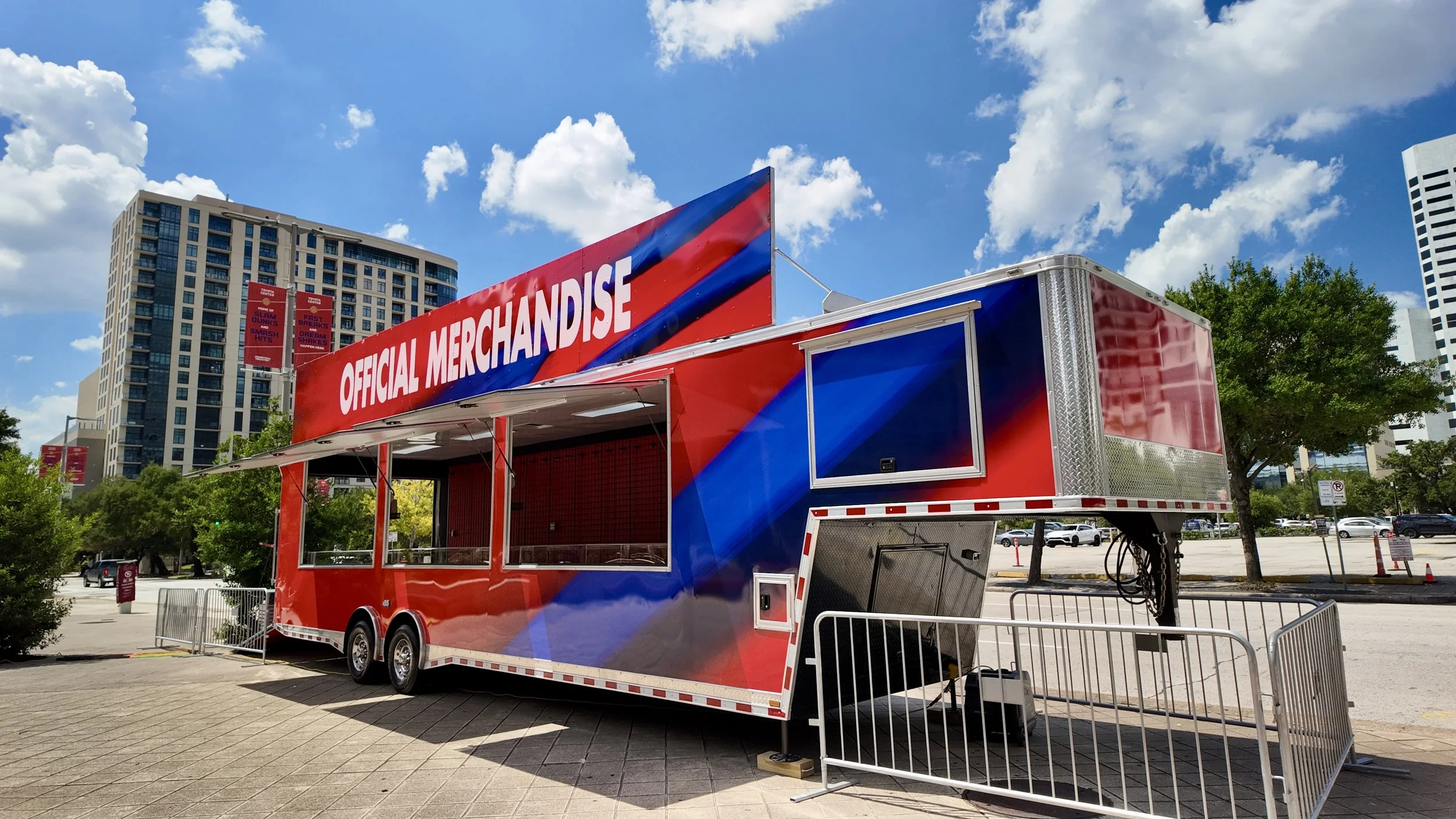 A red and blue mobile merchandise trailer with a large sign reading 'Official Merchandise' parked outdoors in an urban area with high-rise buildings, trees, and cloudy blue sky.