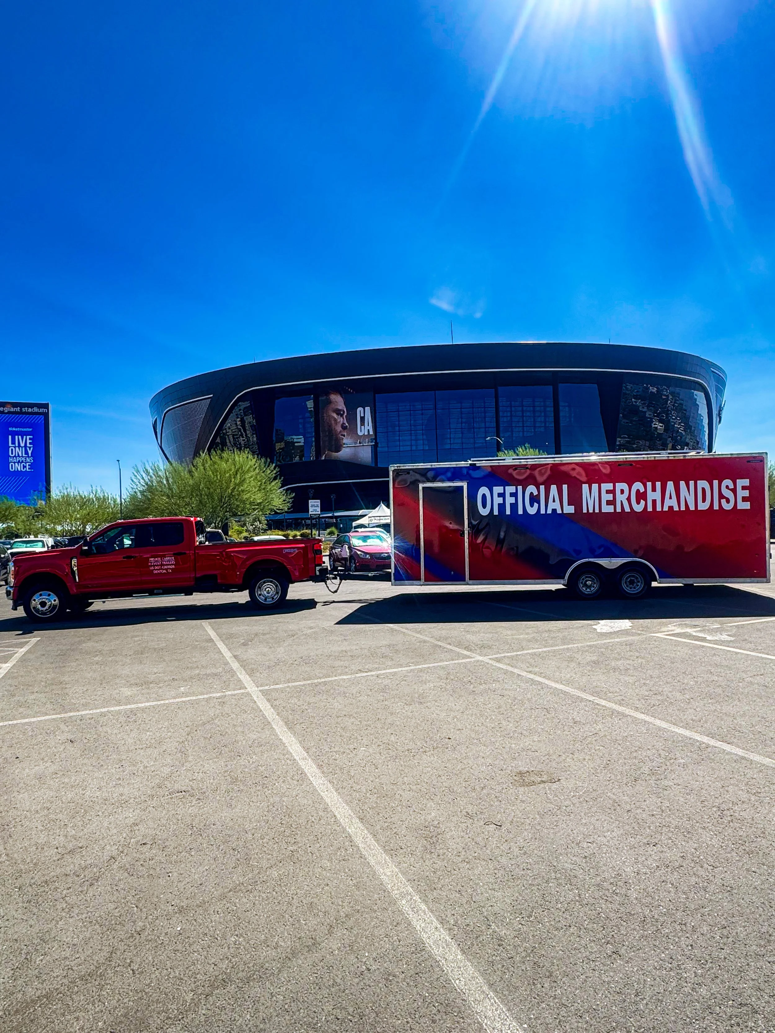 Parking lot with a red pickup truck and a large trailer labeled 'Official Merchandise' parked in front of a modern stadium with a digital screen and a bright sunny sky.