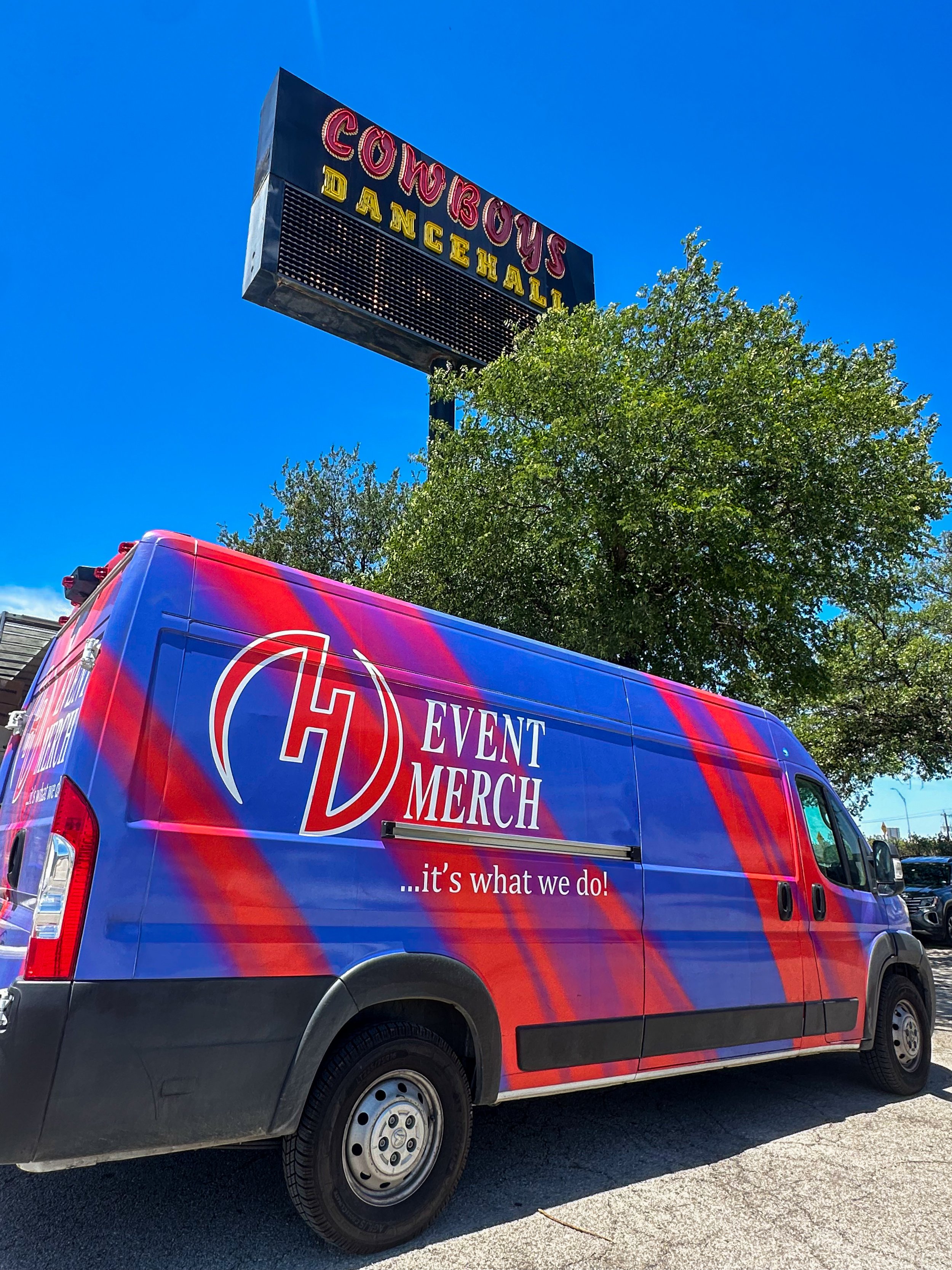 A colorful van with 'Event Merch ...it's what we do!' written on the side parked in front of an electronic marquee sign for Cowboystown Dancehall, under a clear blue sky and trees.