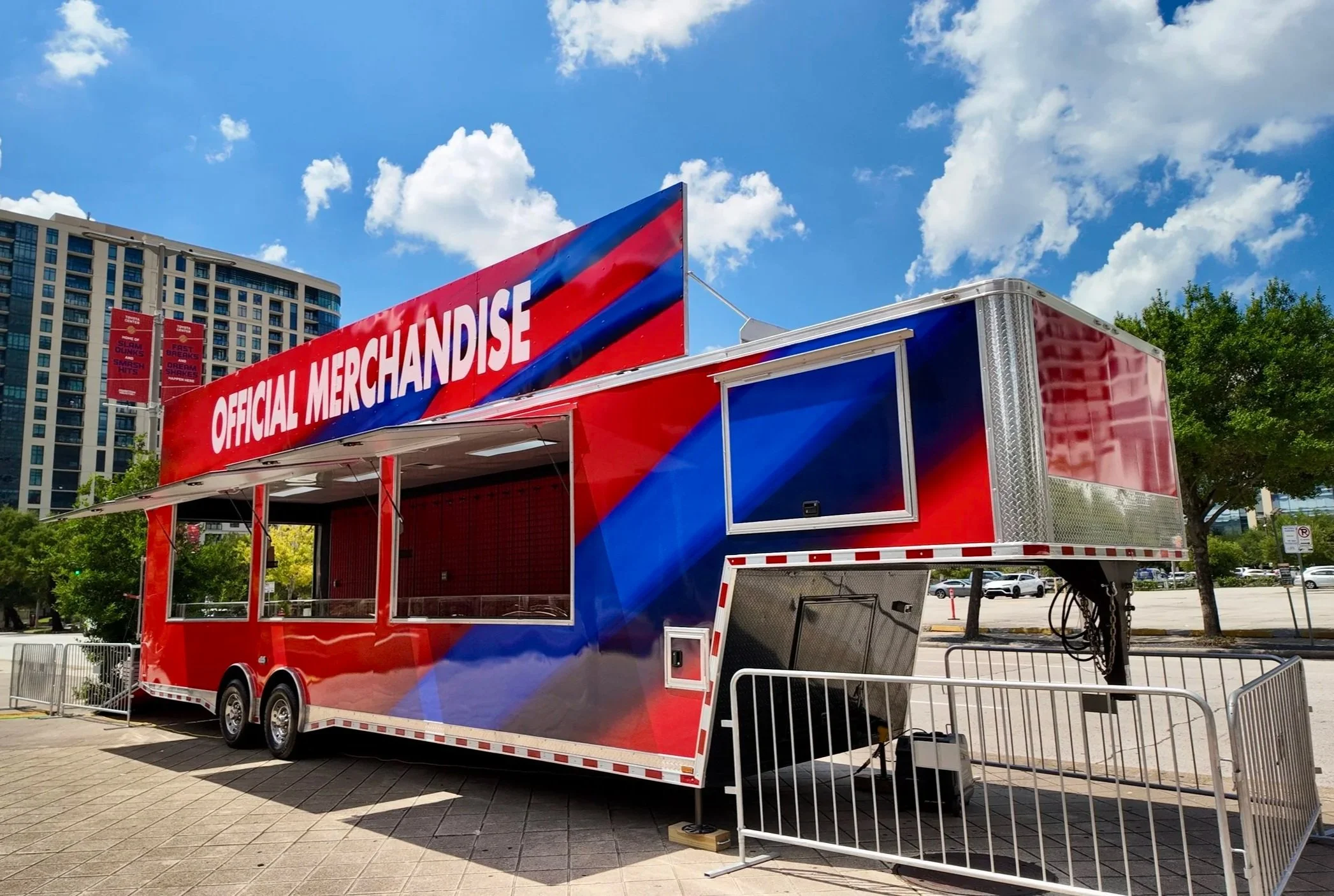 A red, blue, and white official merchandise trailer with open serving windows, parked outdoors on a paved area with nearby trees, a city street, and tall buildings in the background under a partly cloudy sky.