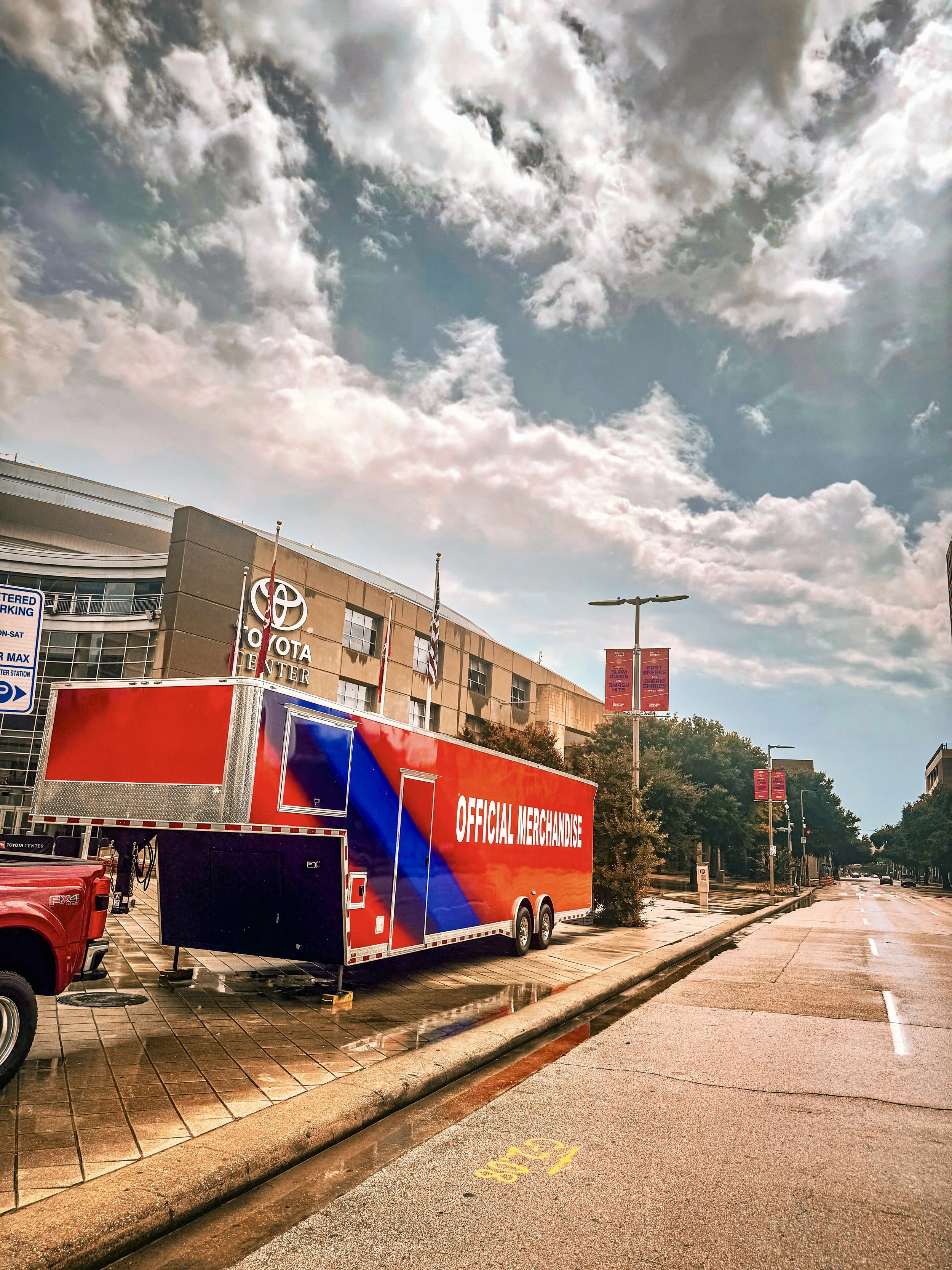 Red trailer with white and blue graphics and the words "Official Merchandise" parked on a city street near a building with a "Toyota Center" sign, under a partly cloudy sky.