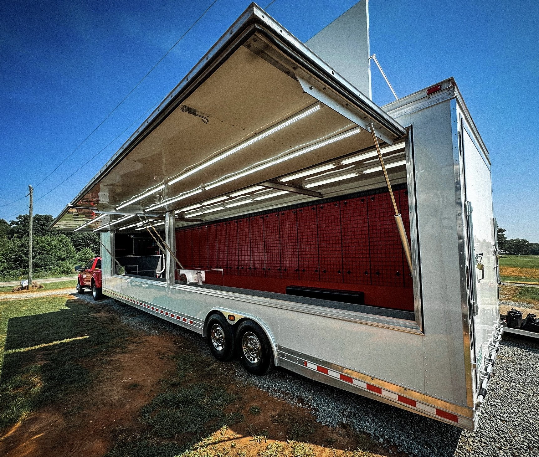 A large mobile food truck with its side open, showing interior fixtures, parked on a grassy area under a clear blue sky.
