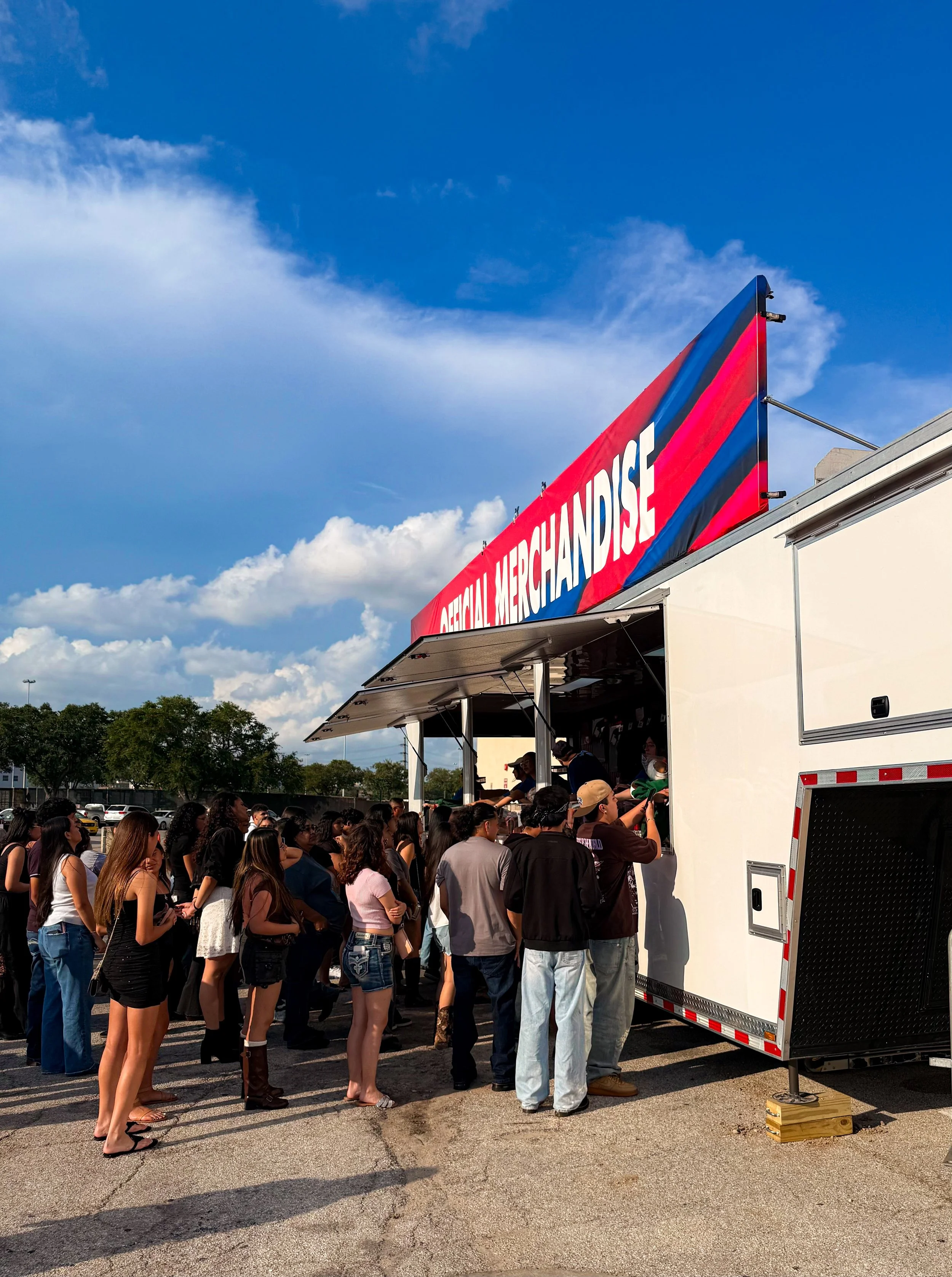 A line of people waiting to buy items from an ATM inside a mobile merchandise store truck with a red, white, and blue sign on top reading 'ACTUAL MERCHANDISE' under a partly cloudy sky.