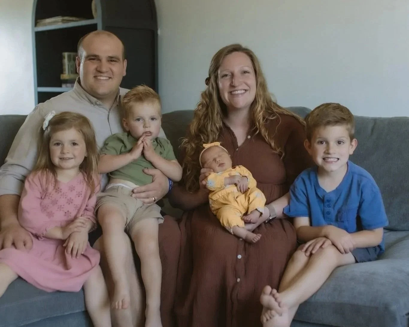 A happy family of five posing outdoors in front of greenery, including a father, mother, two young boys, and a young girl.
