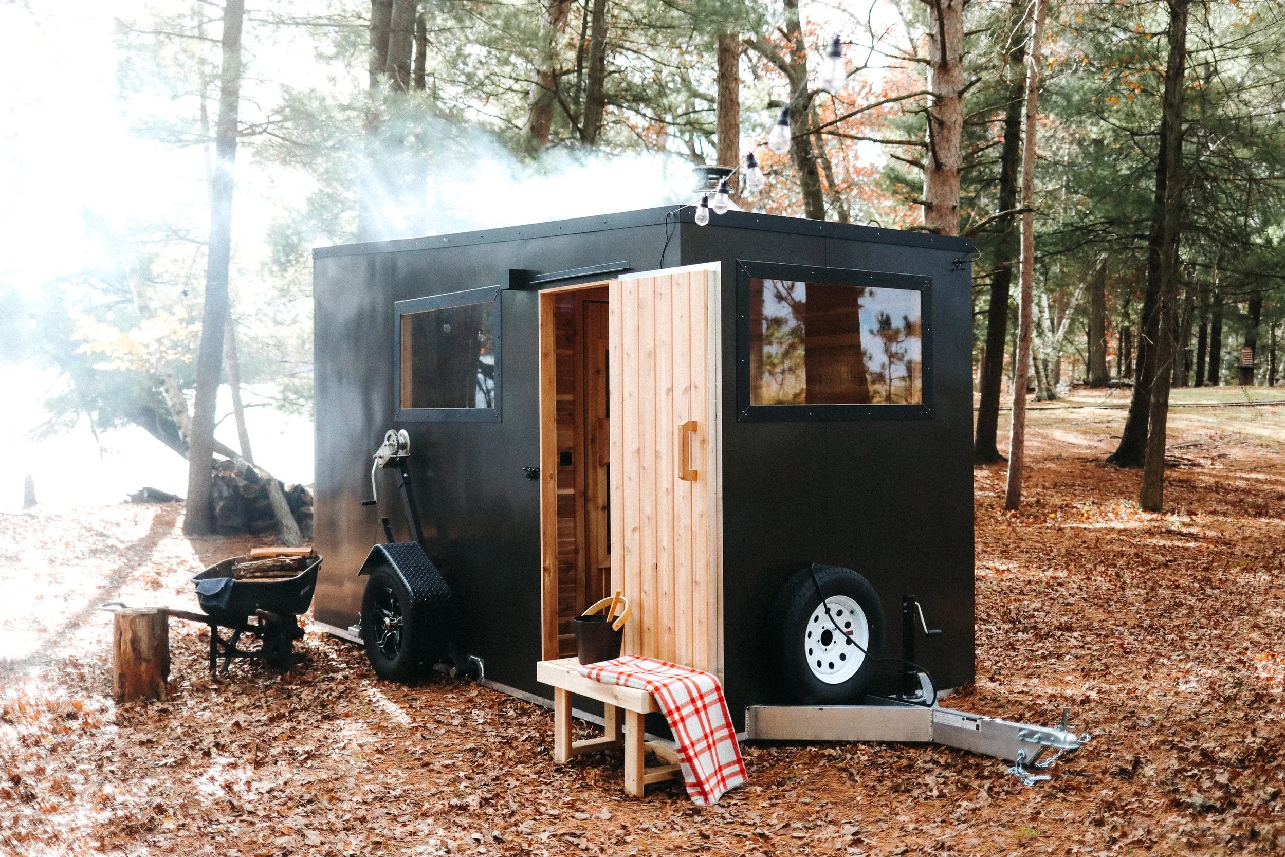 Mobile sauna trailer in a forest setting with an open wooden door, bench, and checkered blanket, surrounded by trees and fallen leaves.