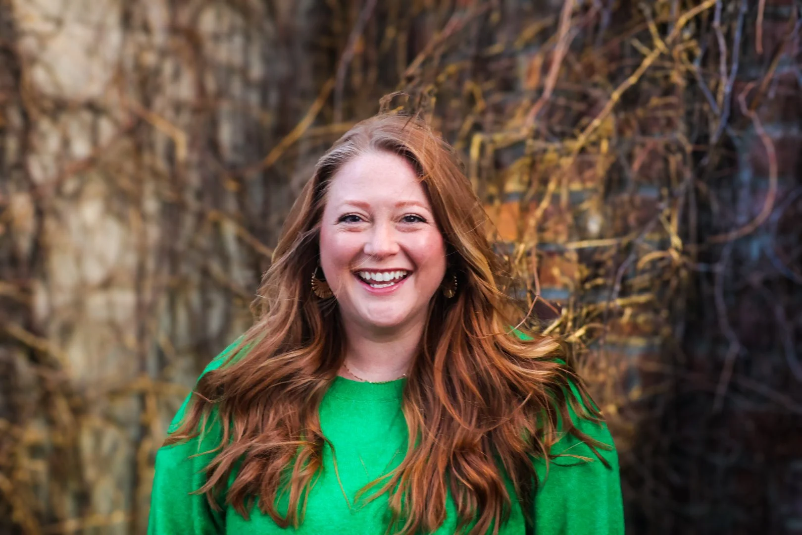 Therapist Chelsea Schrunk Crivello with long red hair wearing a green sweater, smiling in front of a background with vines.