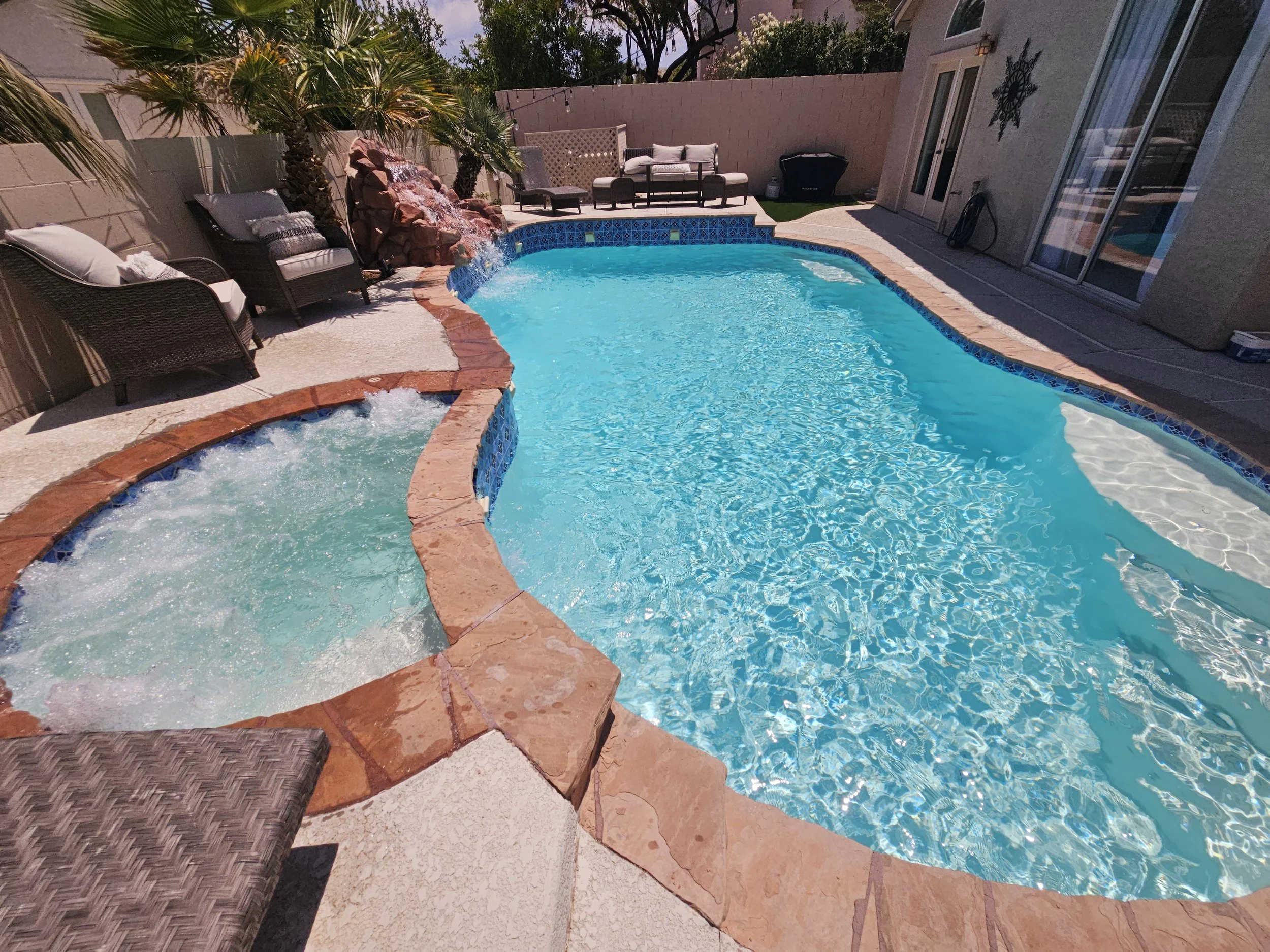 Backyard swimming pool with hot tub, patio furniture, palm trees, rock waterfall, and nearby house with sliding glass doors.