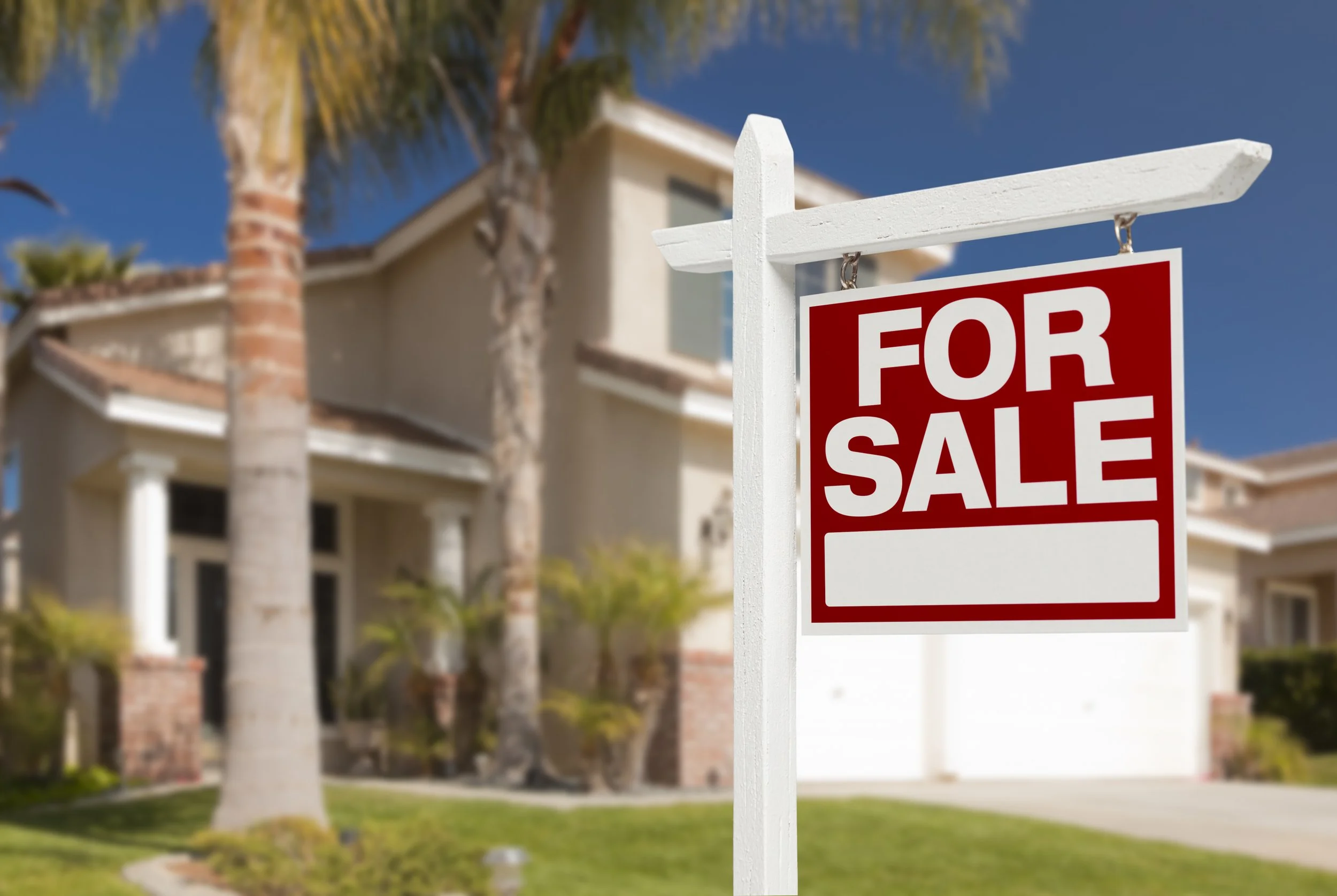 Real estate for sale sign in front of a beige house with palm trees and a blue sky.