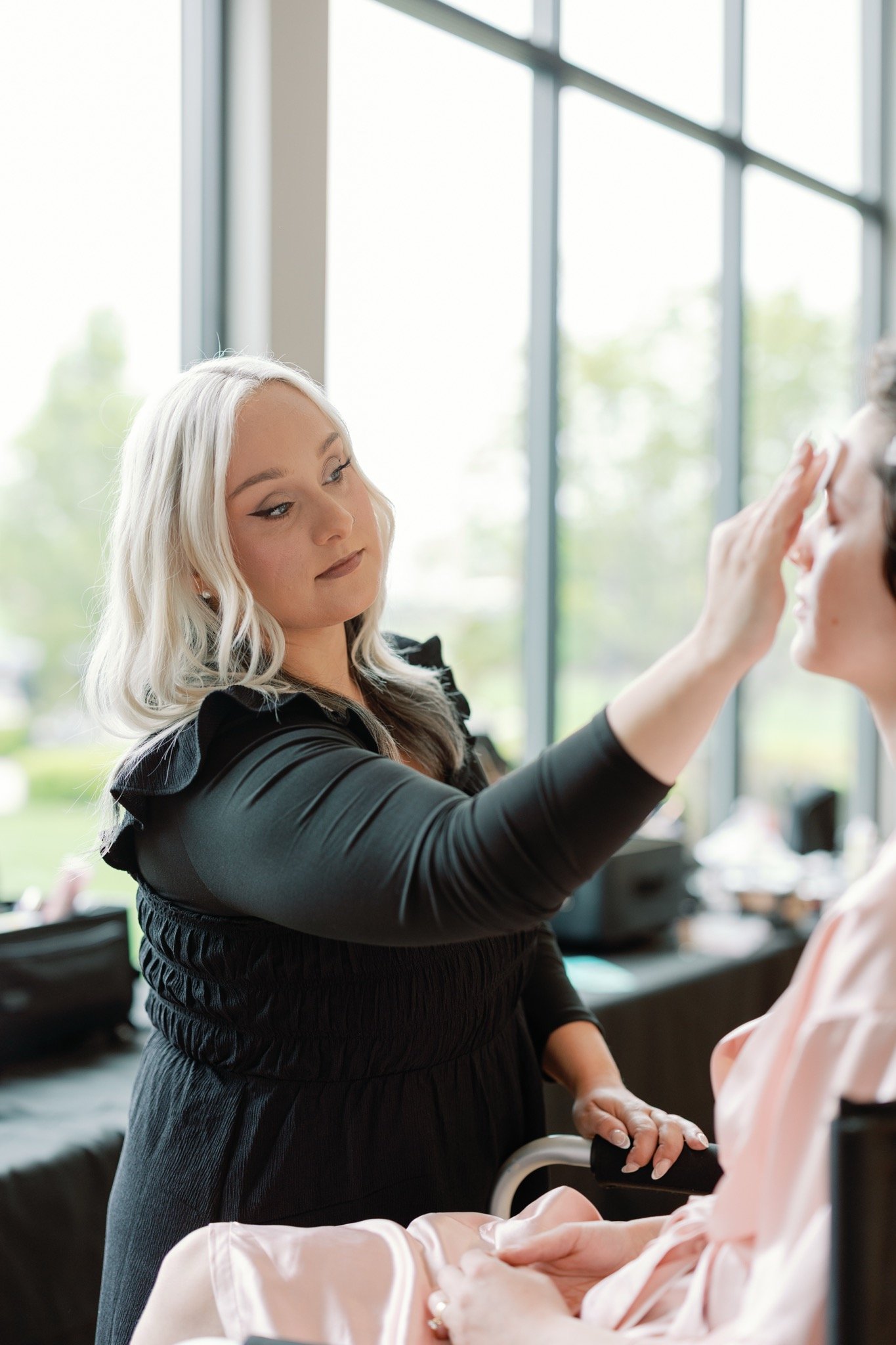 Makeup artist applying makeup to a woman's face in a bright studio with large windows.
