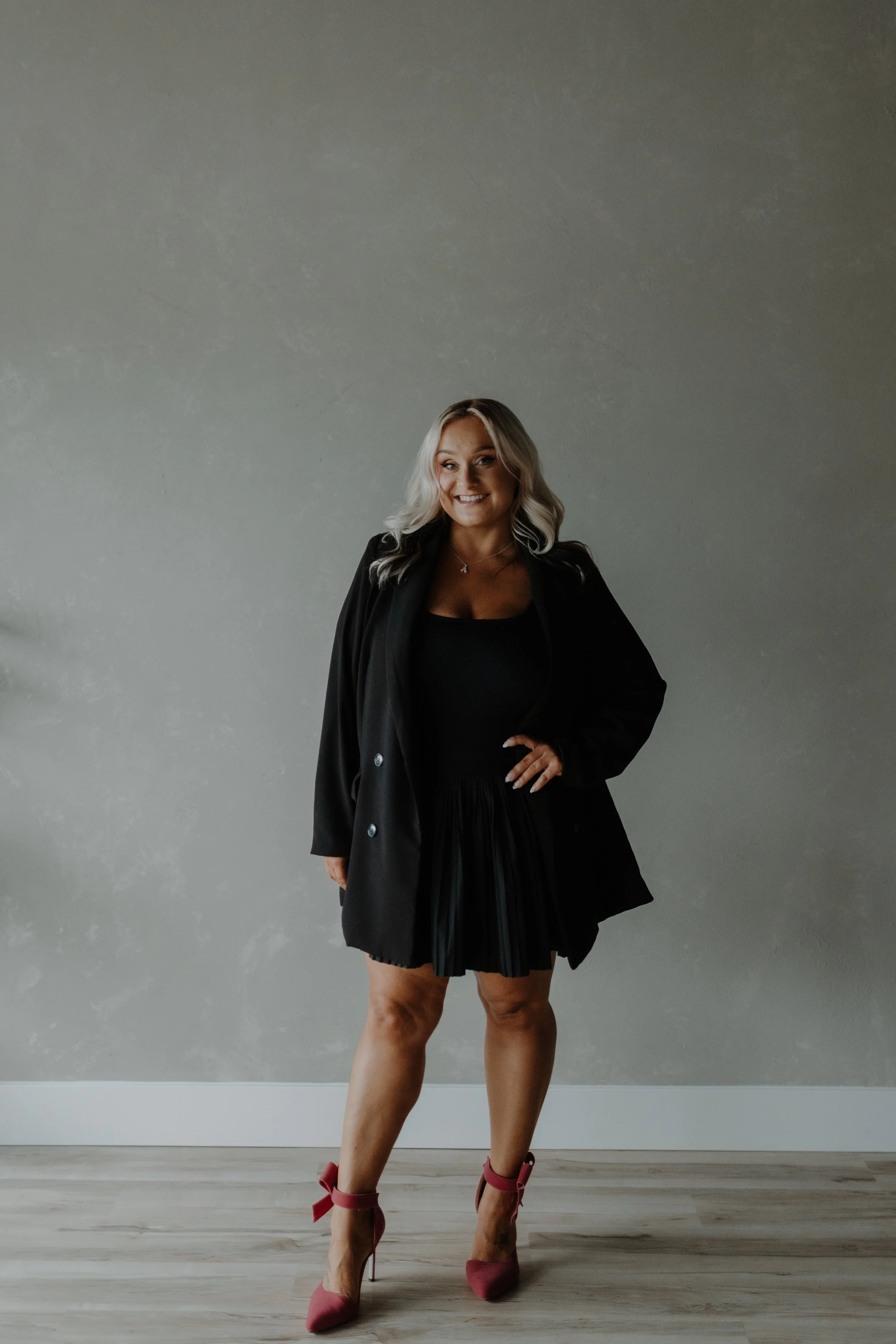 A woman standing indoors against a plain gray wall, wearing a black dress, a black blazer, and pink high-heeled shoes with ankle straps, smiling and posing confidently.
