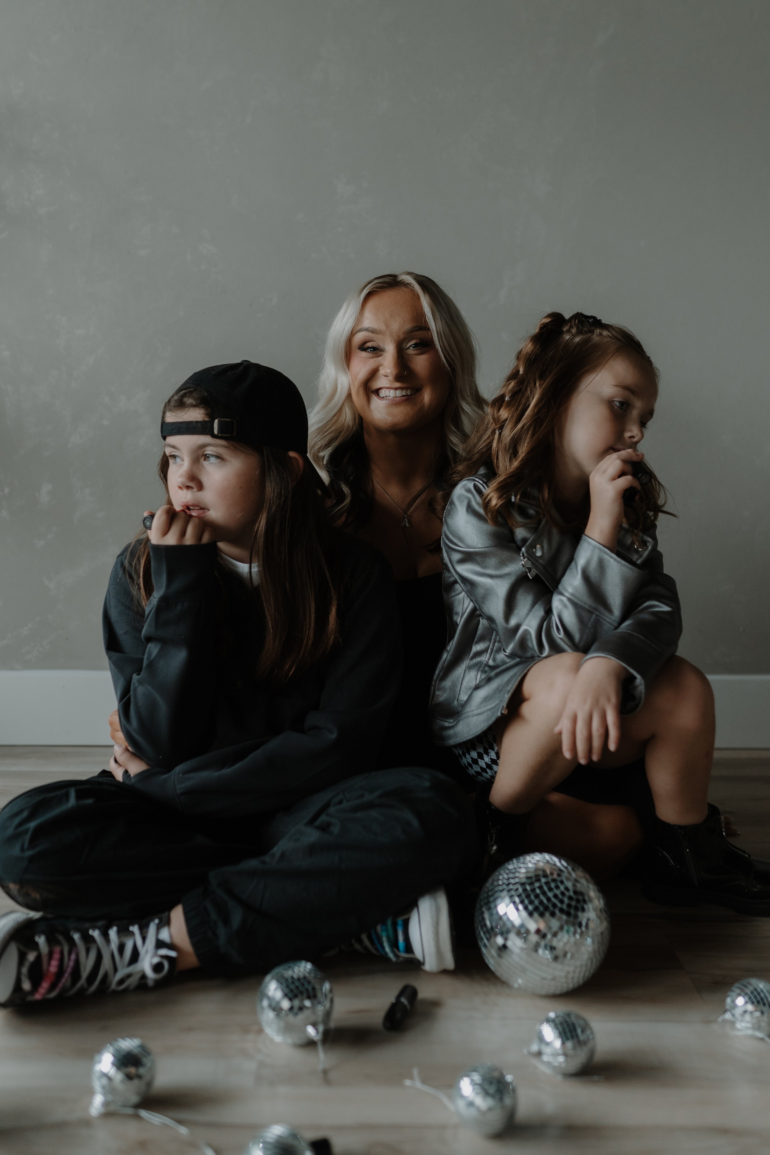 A woman with blonde hair smiling, sitting on the floor with two young girls, one wearing a black baseball cap and the other with wavy hair and a silver jacket, surrounded by small disco balls.
