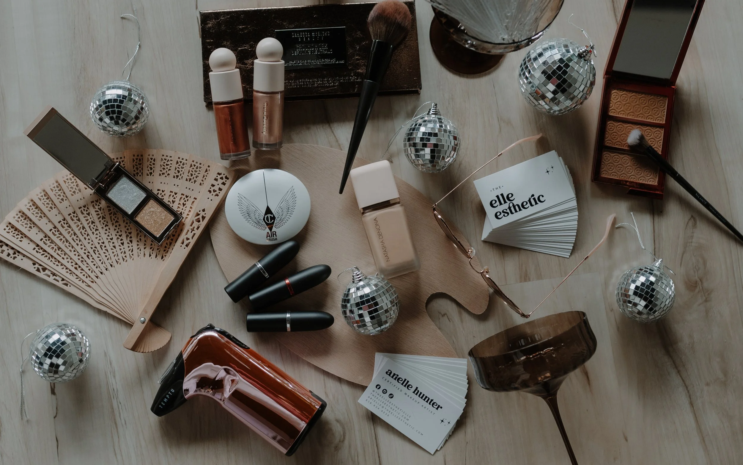 A flat lay of beauty and makeup products, including lipsticks, foundation, eyeshadow, mascara, a makeup brush, sunglasses, decorative disco balls, a fan, a business card, a container, and a glass, arranged on a wooden surface.
