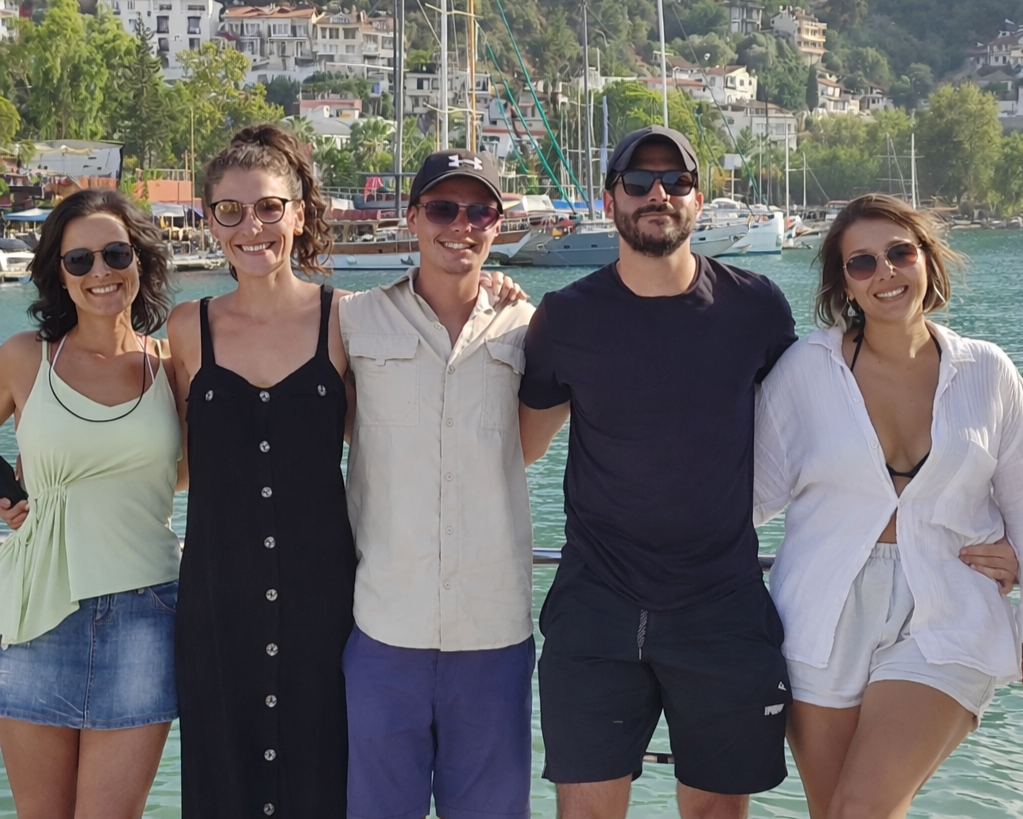 Group of five friends standing arm-in-arm on a boat dock with boats and hillside homes in the background. They are smiling and wearing sunglasses, dressed in summer clothing.