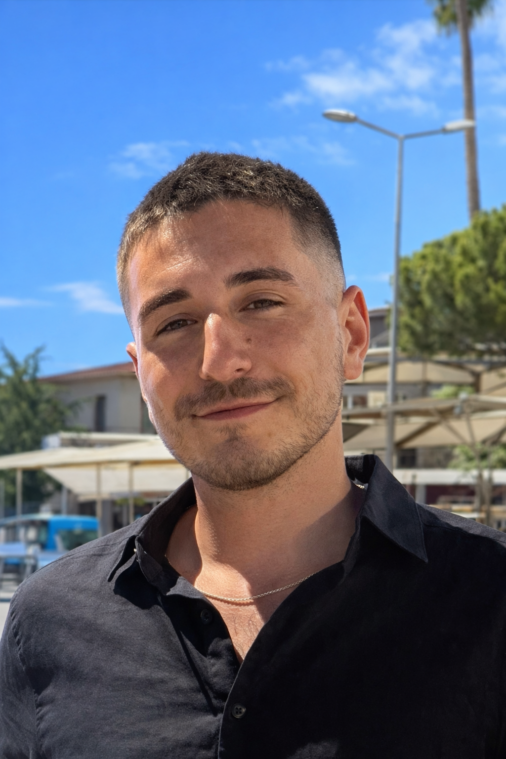 A young man with short brown hair and a slight beard, smiling outdoors on a sunny day with a blue sky, trees, and building structures in the background.