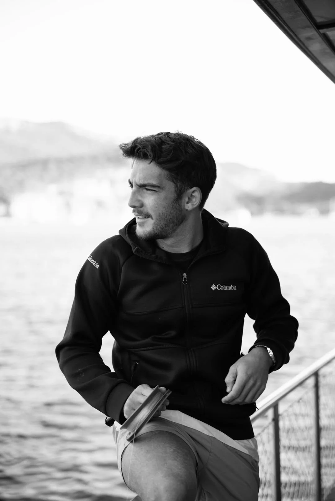 A young man with dark hair and a beard, wearing a Columbia jacket and shorts, stands on a boat deck by the water, looking to the side with a thoughtful expression.