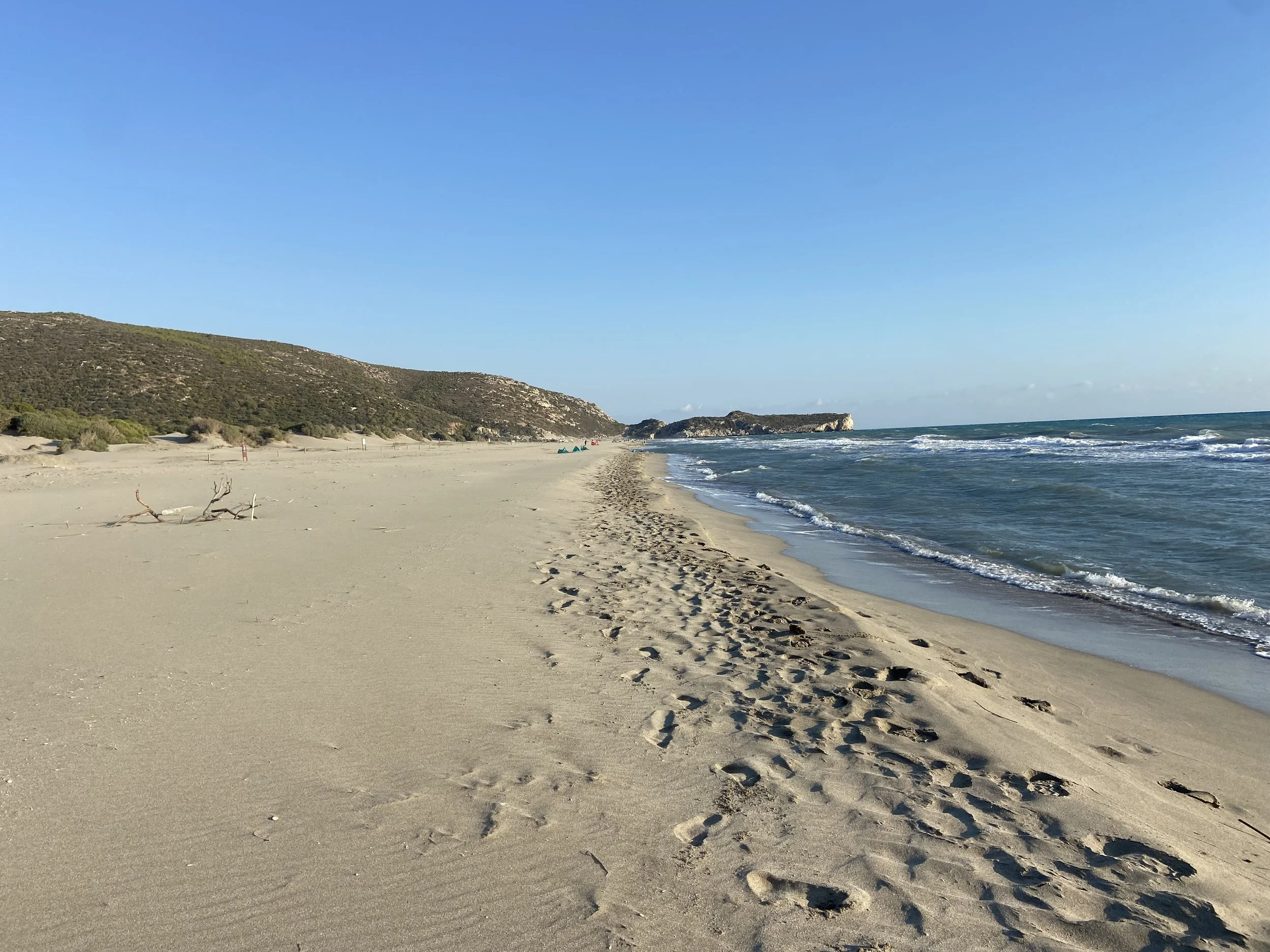 Empty sandy beach with footprints, small driftwood, distant hills, and a rocky outcrop in the ocean under a clear blue sky.