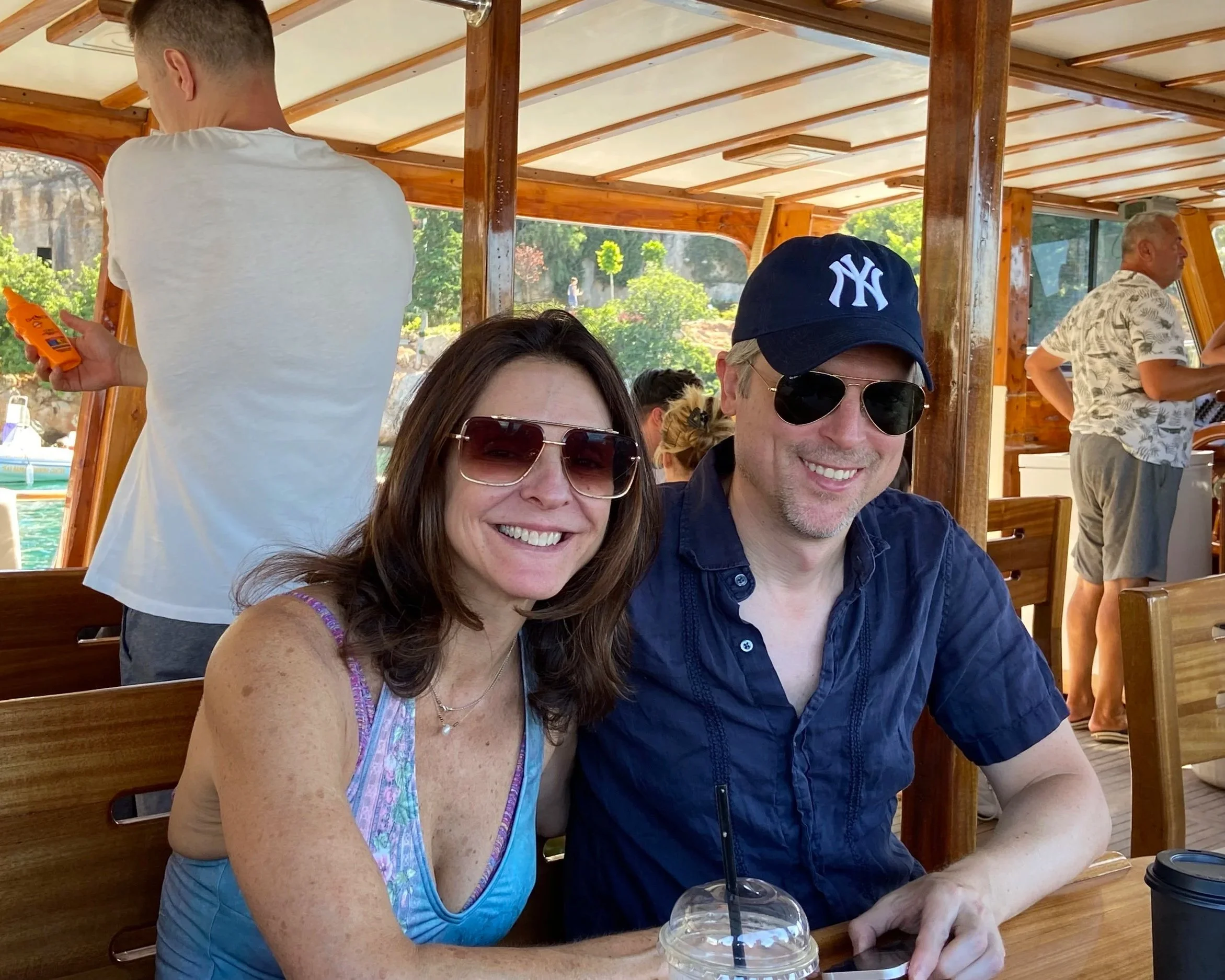 Smiling woman and man sitting at a table on a boat or indoor patio, with a scenic outdoor view of greenery and water in the background, wearing sunglasses and casual summer clothes.