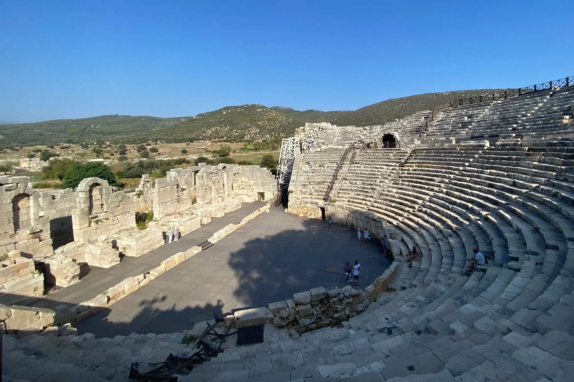 Ancient Greek theater with stone seating and stage, with hills and clear blue sky in the background.