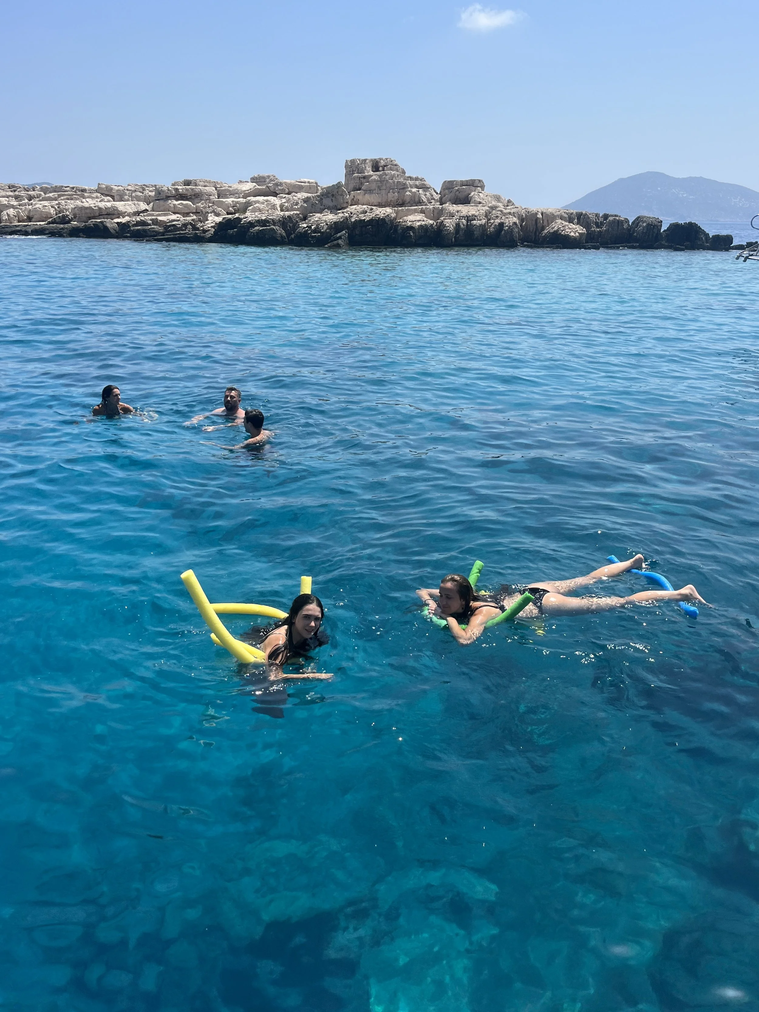 Group of people swimming and floating with pool noodles in a clear blue body of water near rocky shoreline and hills in the background.