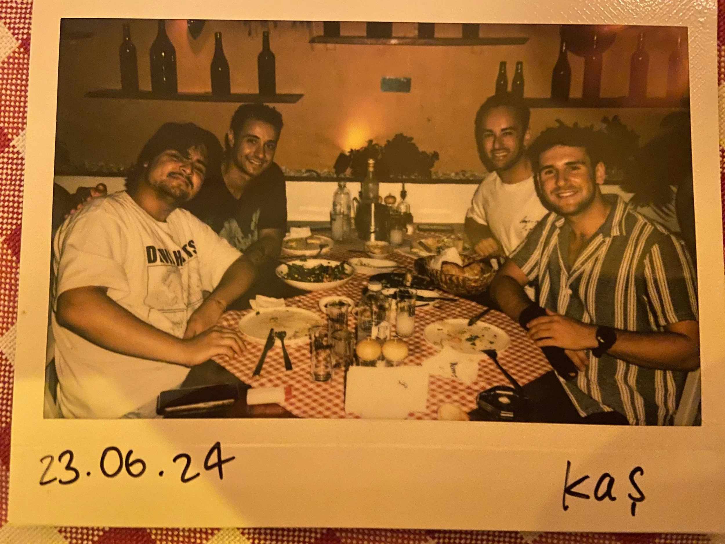 Group of five young men enjoying a meal together at a restaurant with a checked tablecloth and bottles on shelves in the background.