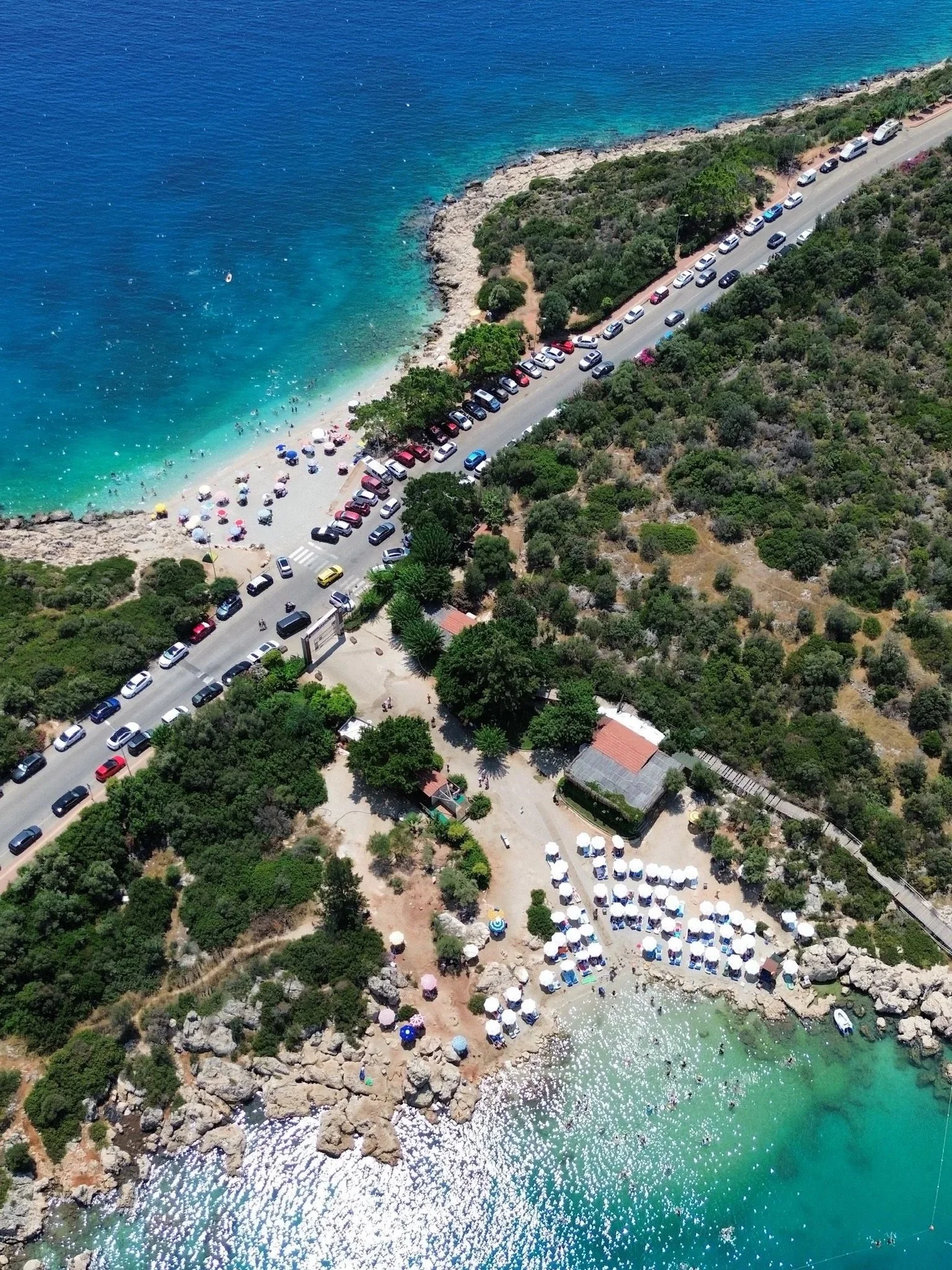 Aerial view of a coastal beach with clear blue water, sandy shoreline with umbrellas, and a parking lot with many cars beside lush green trees.