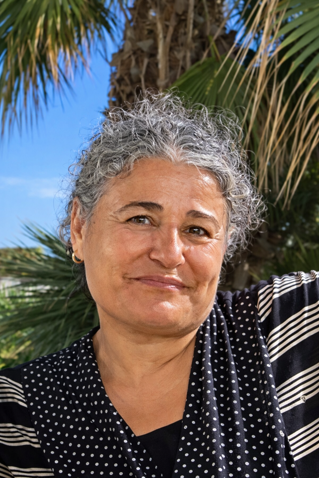 A woman with curly gray hair smiling outdoors with palm trees and a blue sky in the background.
