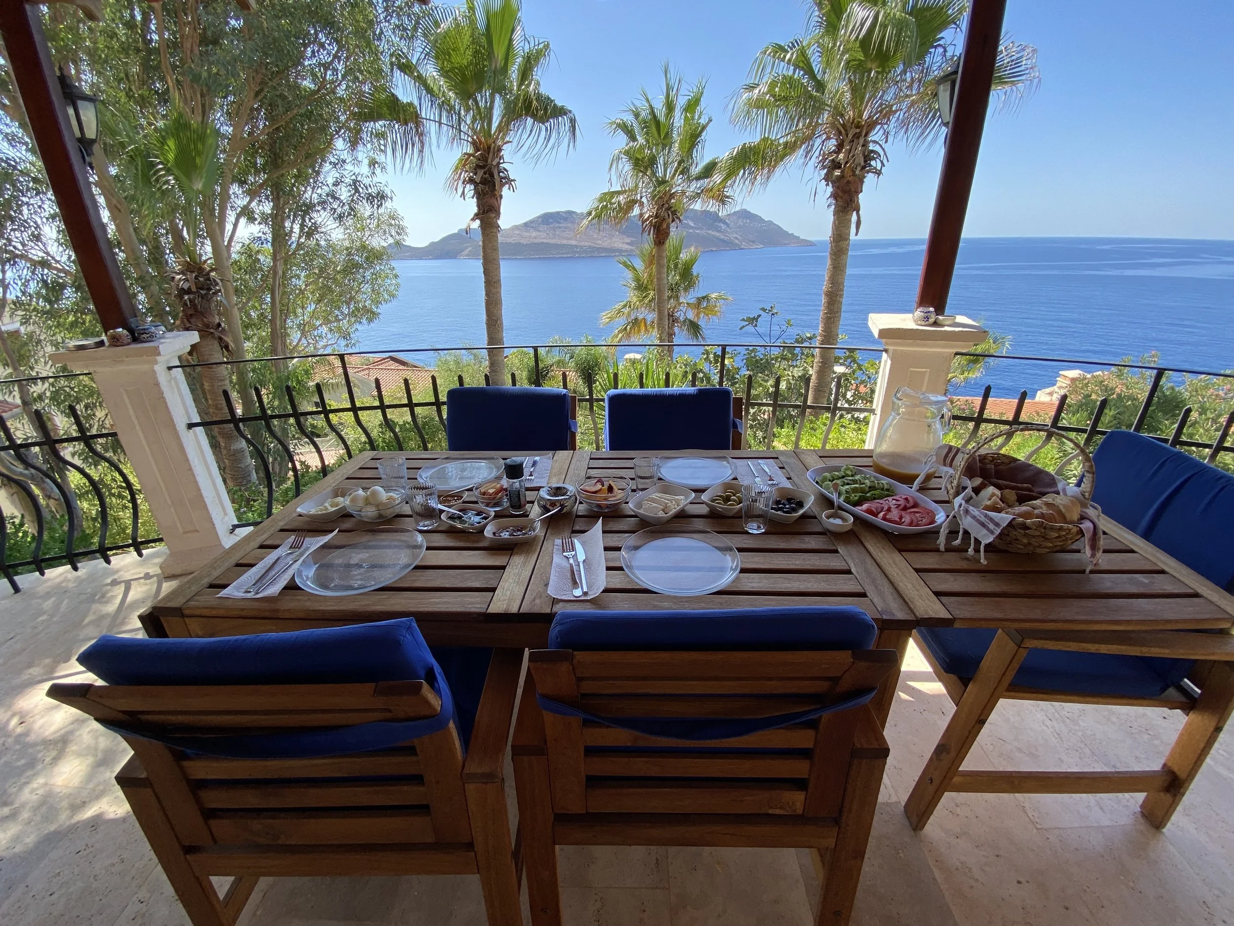 A wooden outdoor dining table set for breakfast with dishes, bread basket, and fruit, overlooking a seaside view with palm trees and mountains.