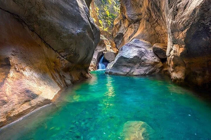 A narrow canyon with smooth, towering rock walls and a small waterfall flowing into a clear, turquoise pool of water.