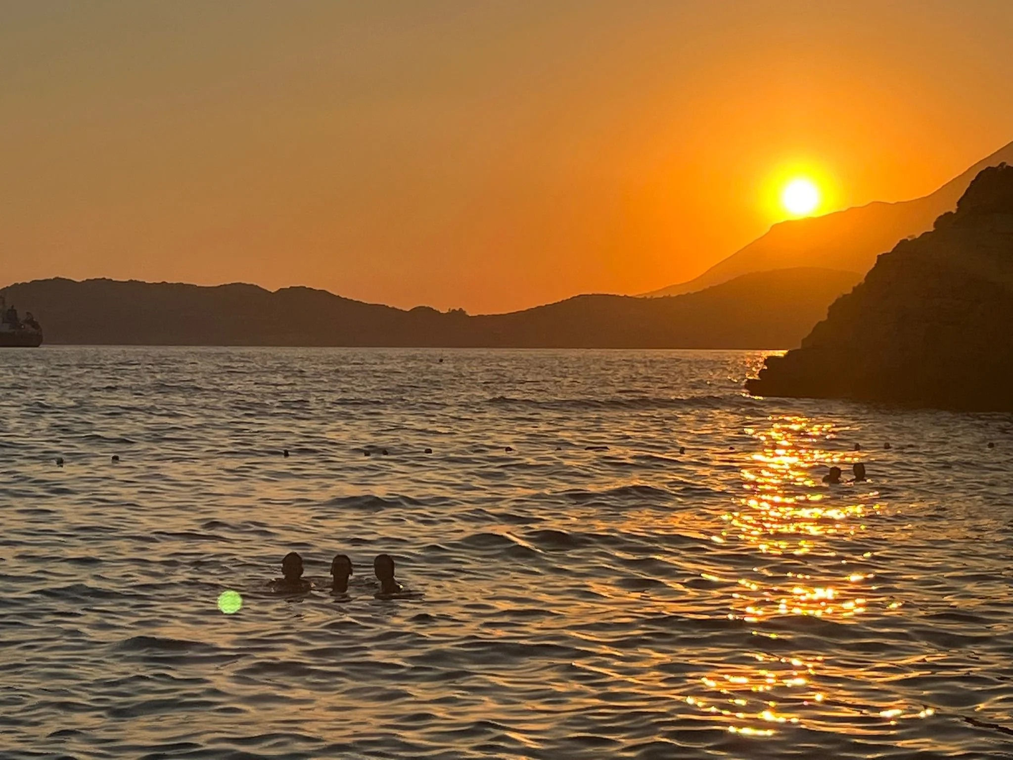 Sunset over the ocean with three people swimming in the water in the foreground, a boat on the left, and hills in the background.