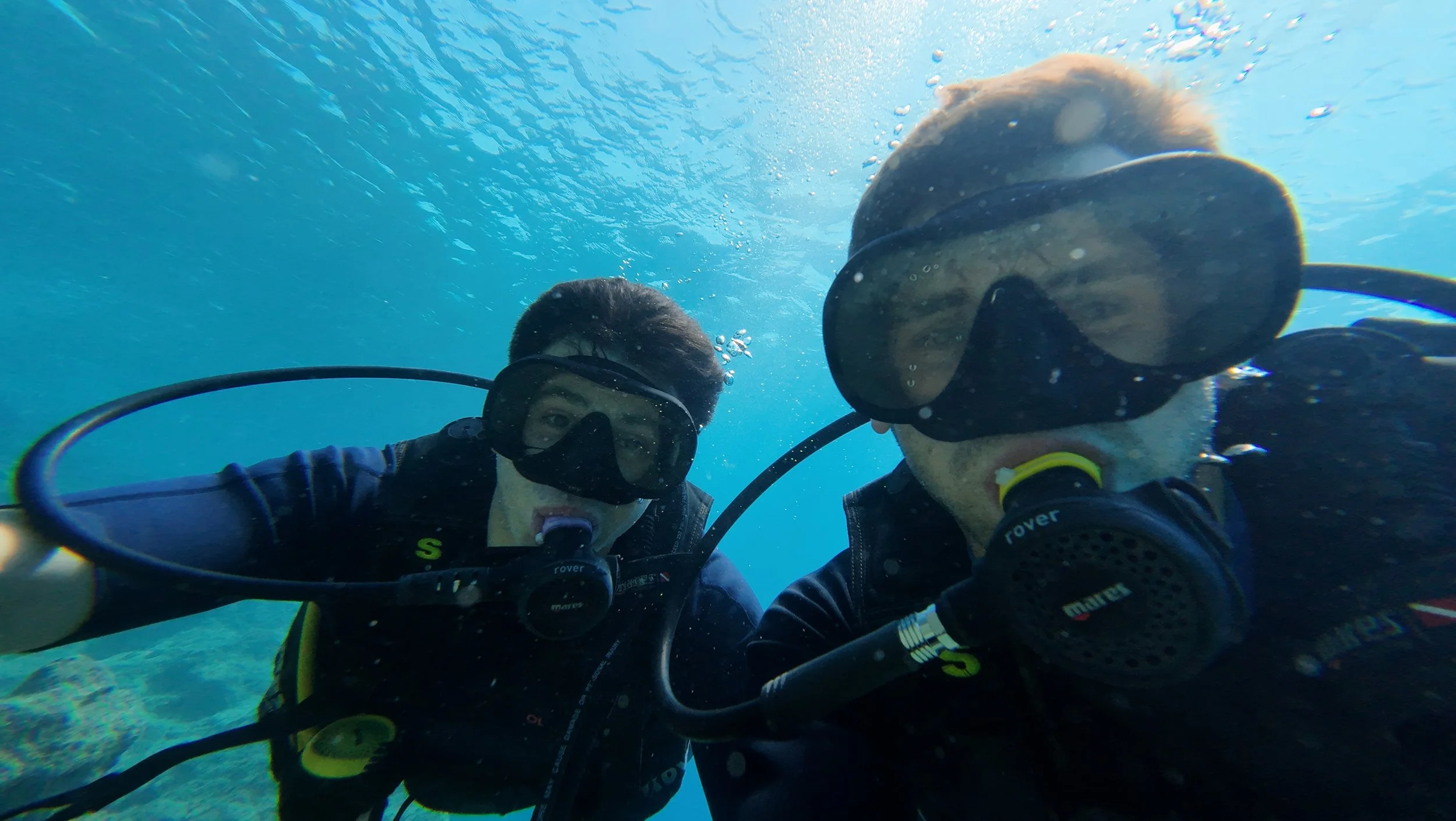 Two people scuba diving underwater, wearing masks and breathing through regulators, taking a selfie.