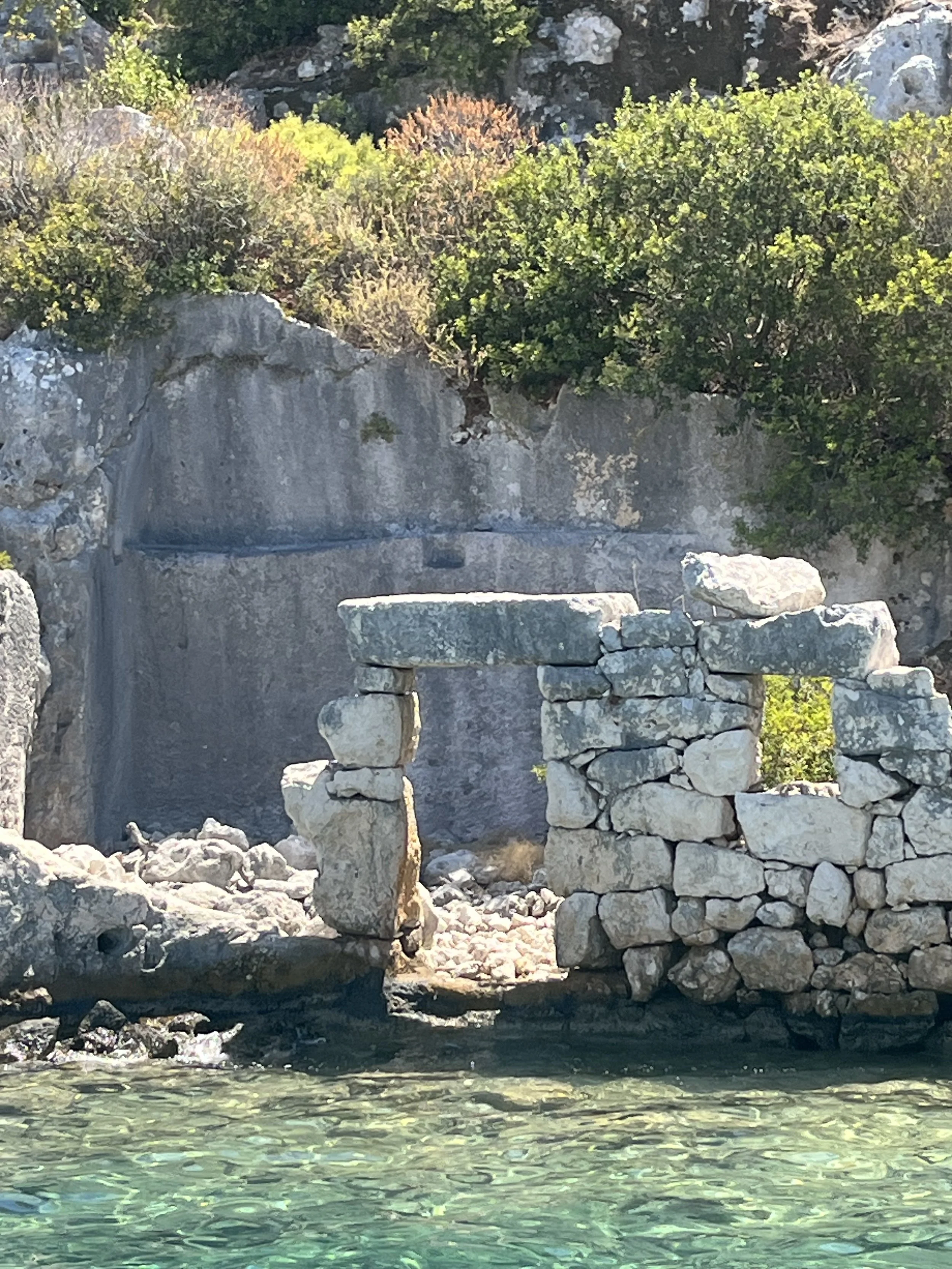 A rustic stone structure on the seaside, with a stone arch and window, surrounded by rocky terrain and lush green bushes and trees in the background.