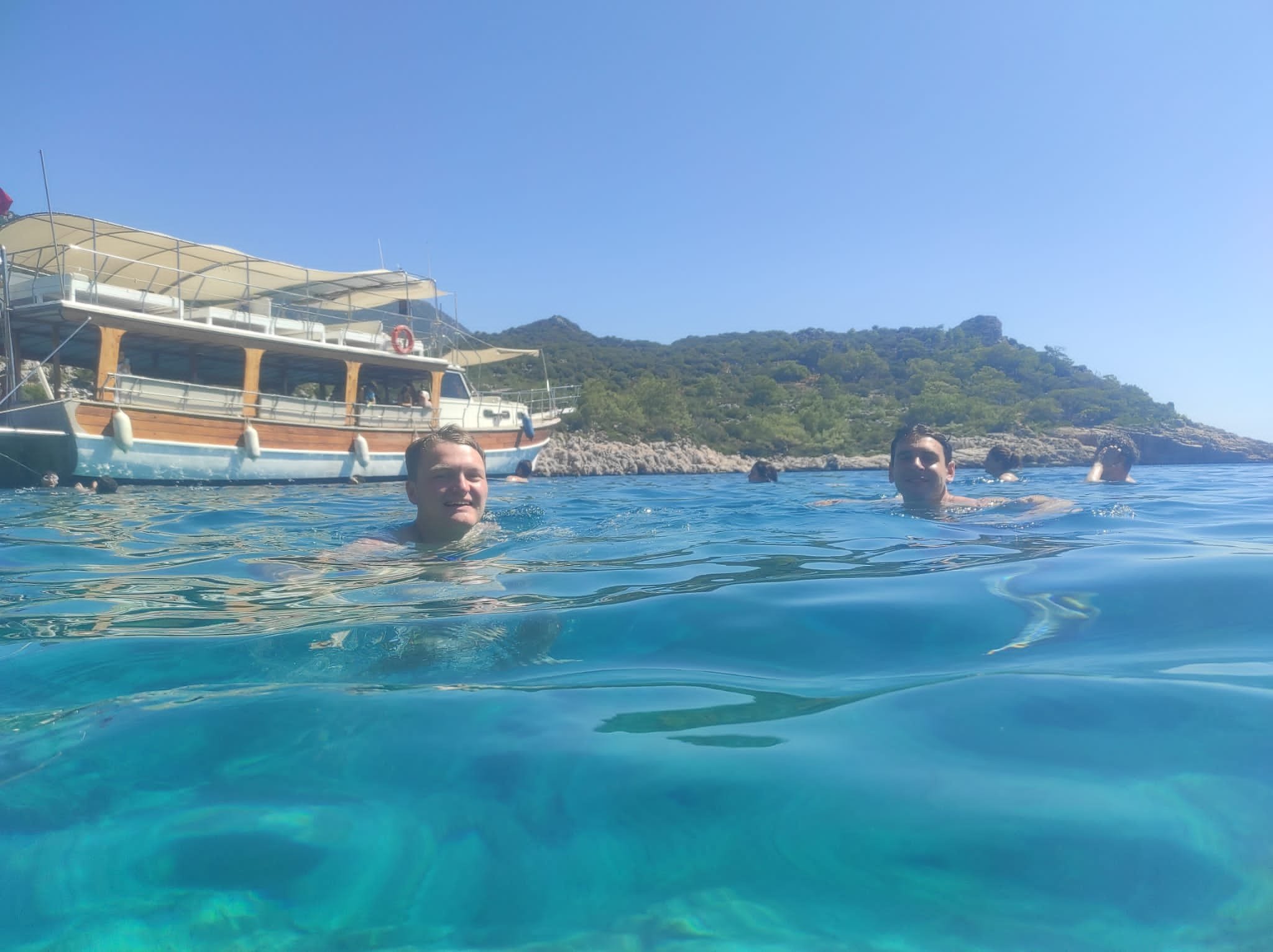 Two people swimming in clear blue water near a boat and rocky shoreline.