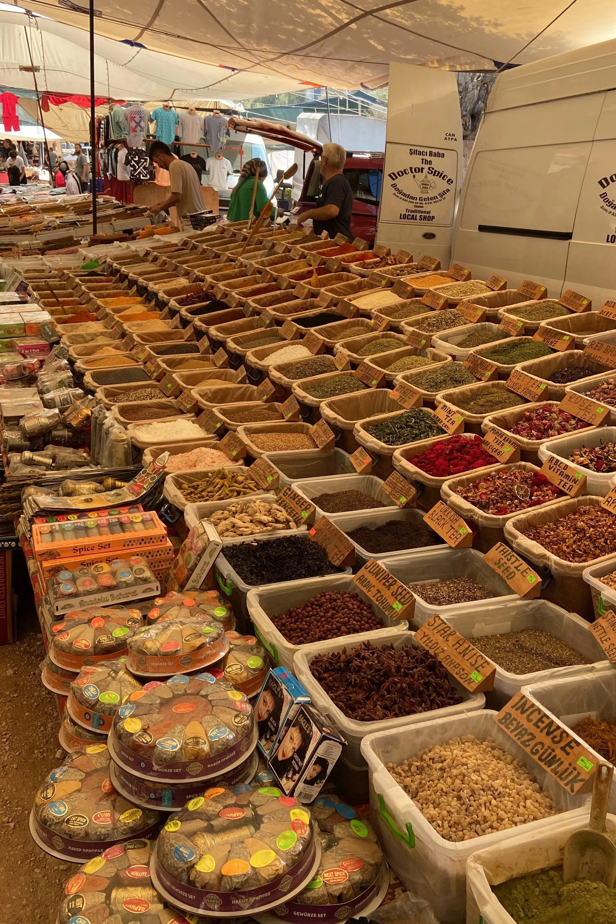 A market stall with a variety of spices and herbs arranged in large containers and jars, with some packaged spice sets in front. Several people are browsing and shopping in the background under a canopy.