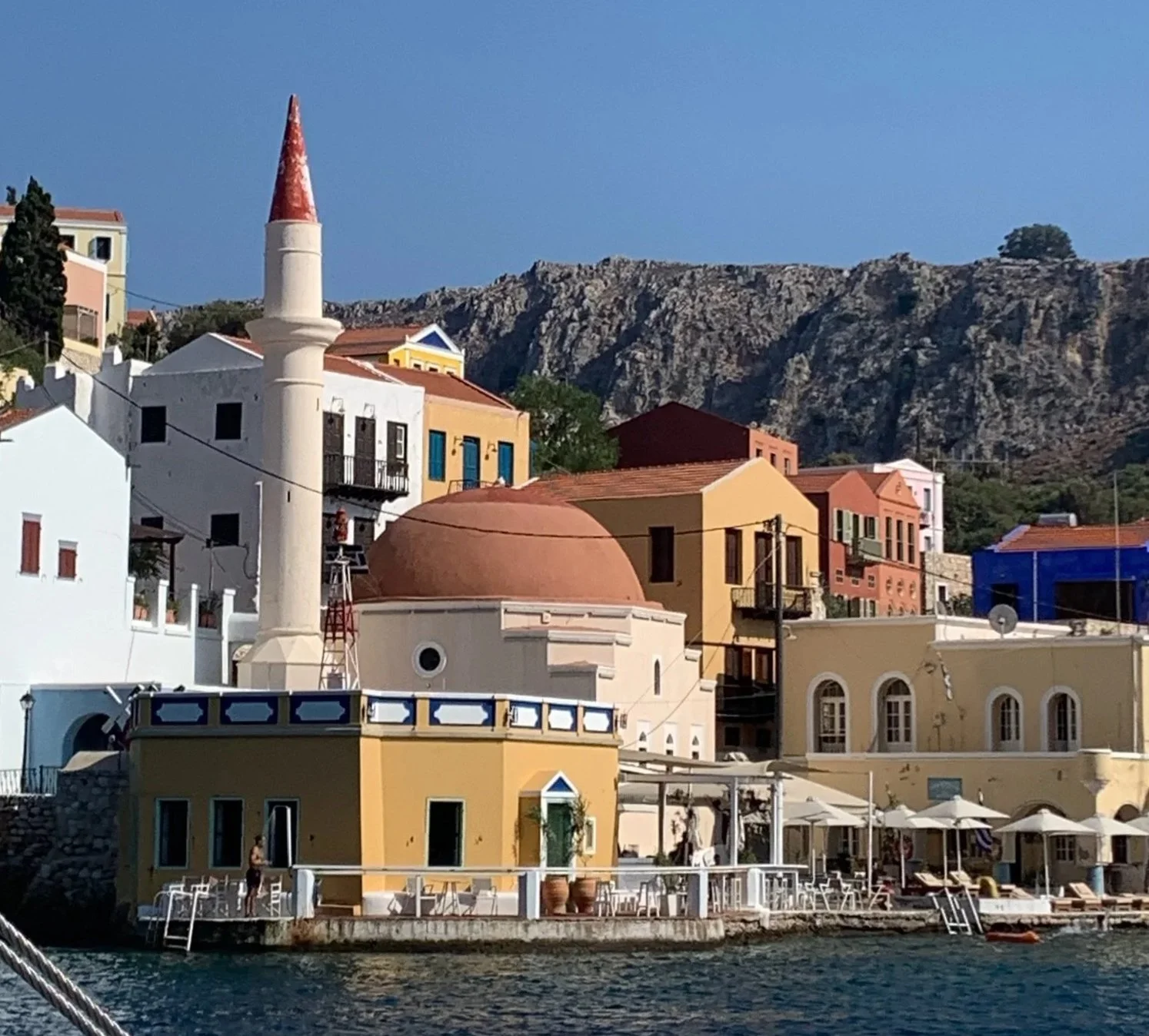 Colorful buildings with a mountain in the background along a waterfront, including a small white and yellow structure with outdoor seating and umbrellas.