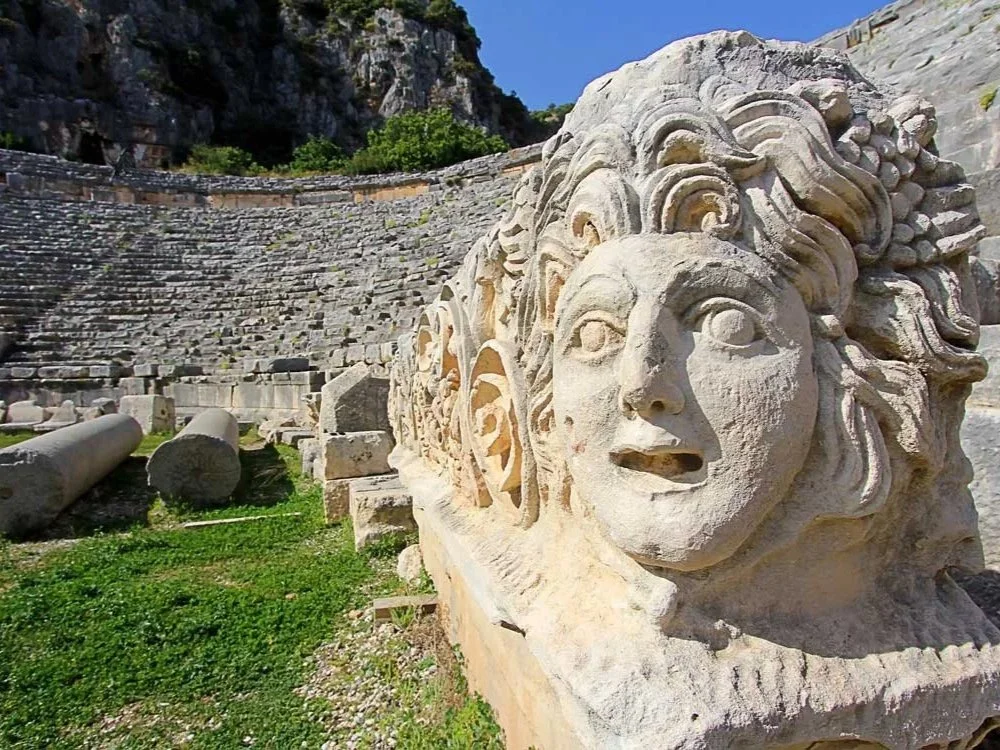 Ancient Greek stone theater with a carved stone lion's head sculpture in the foreground, with stairs and rocky hills in the background.