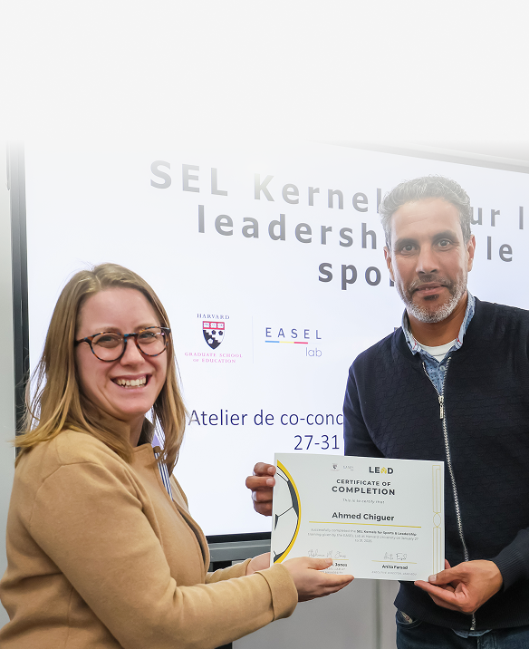 A woman with glasses smiling and a man holding a certificate while standing in front of a presentation screen about leadership and sports.