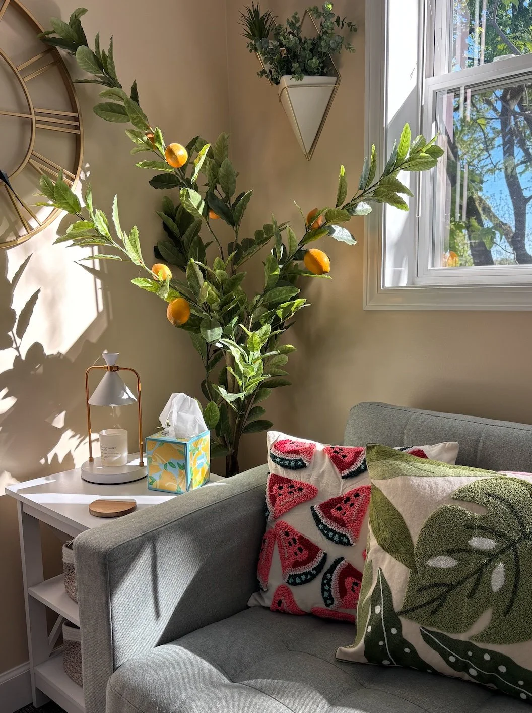 A cozy living room corner with a light gray sofa adorned with colorful watermelon and leafy patterned pillows. Next to the sofa, there is a white side table with a decorative lamp, tissue box, and wooden coaster. A tall lemon tree with green leaves and yellow lemons stands behind the sofa, near an open window letting in natural sunlight. There are also wall-mounted planters and a large wall clock.