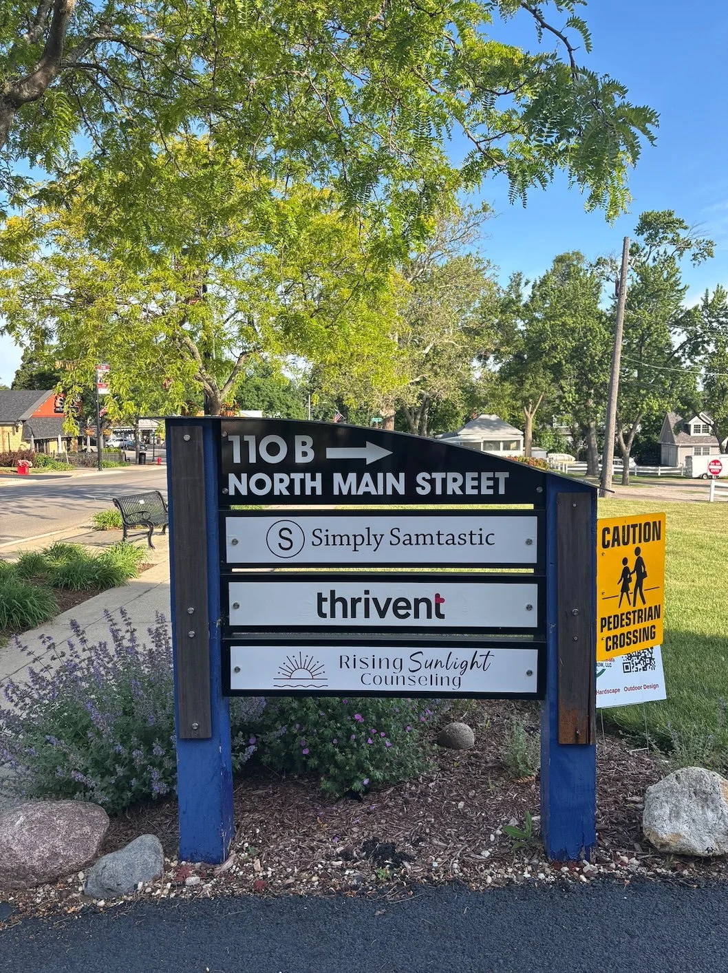 Signboard on North Main Street indicating nearby businesses: Simply Samtastic, Thrivent, Rising Sunlight Counseling; yellow pedestrian crossing caution sign; trees, sidewalk, and houses in the background.