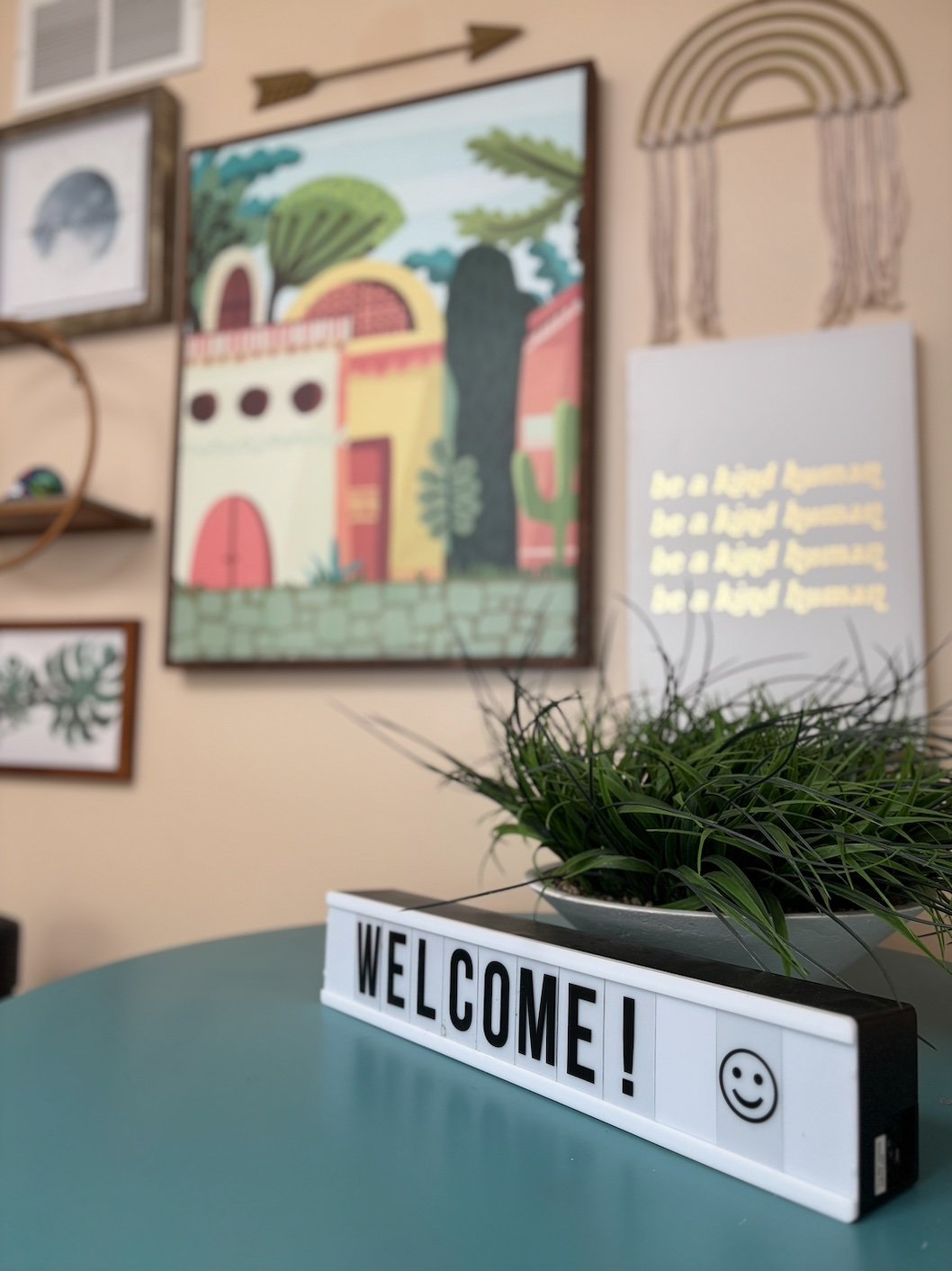 A decorative table with a box sign saying 'WELCOME!' and a smiley face, with a potted plant on top. In the background, there are framed artwork and a marquee styled wall hanging.